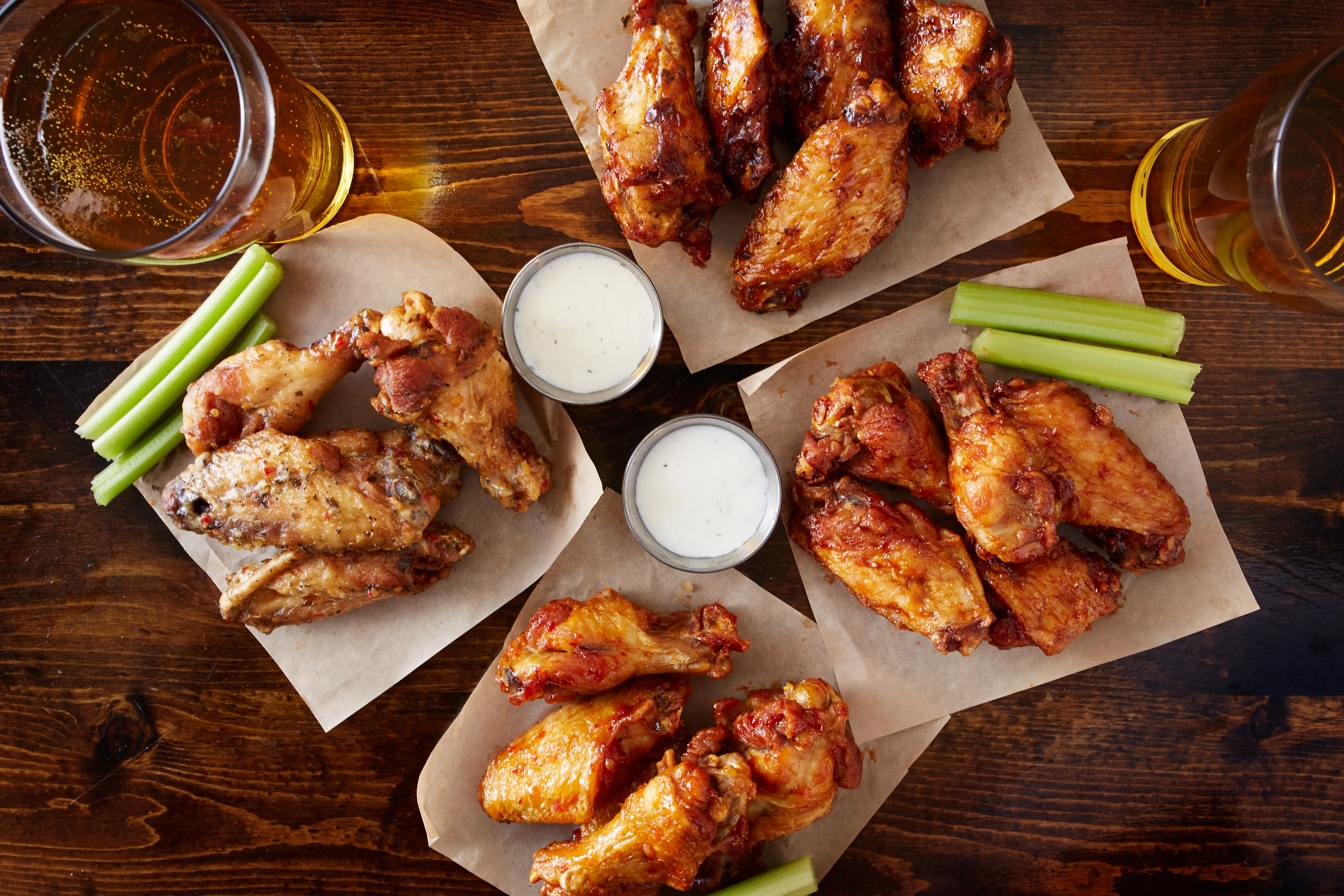 Overhead view of four different flavored chicken wings with celery, sauce and beer on a wood table