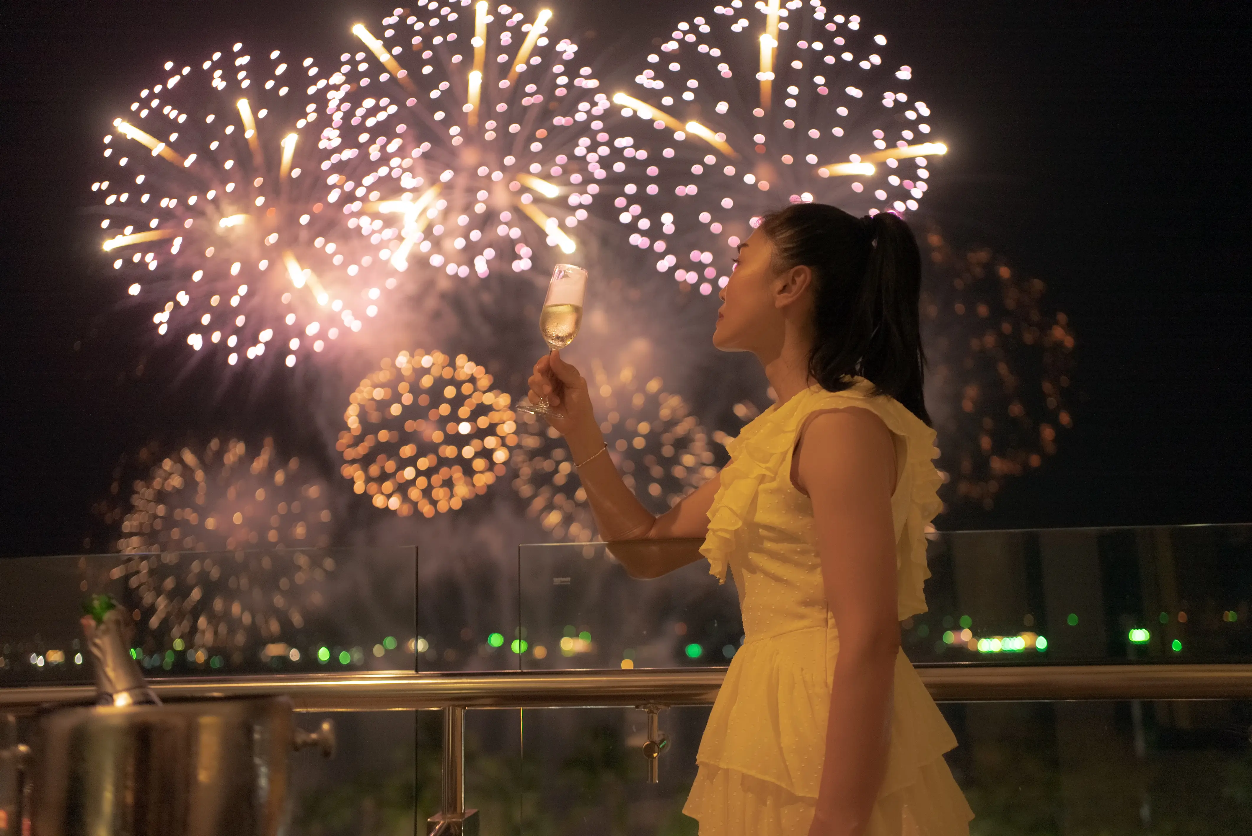 Image of a woman on a ship deck, holding a glass of champagne, and watching the overhead nighttime fireworks.