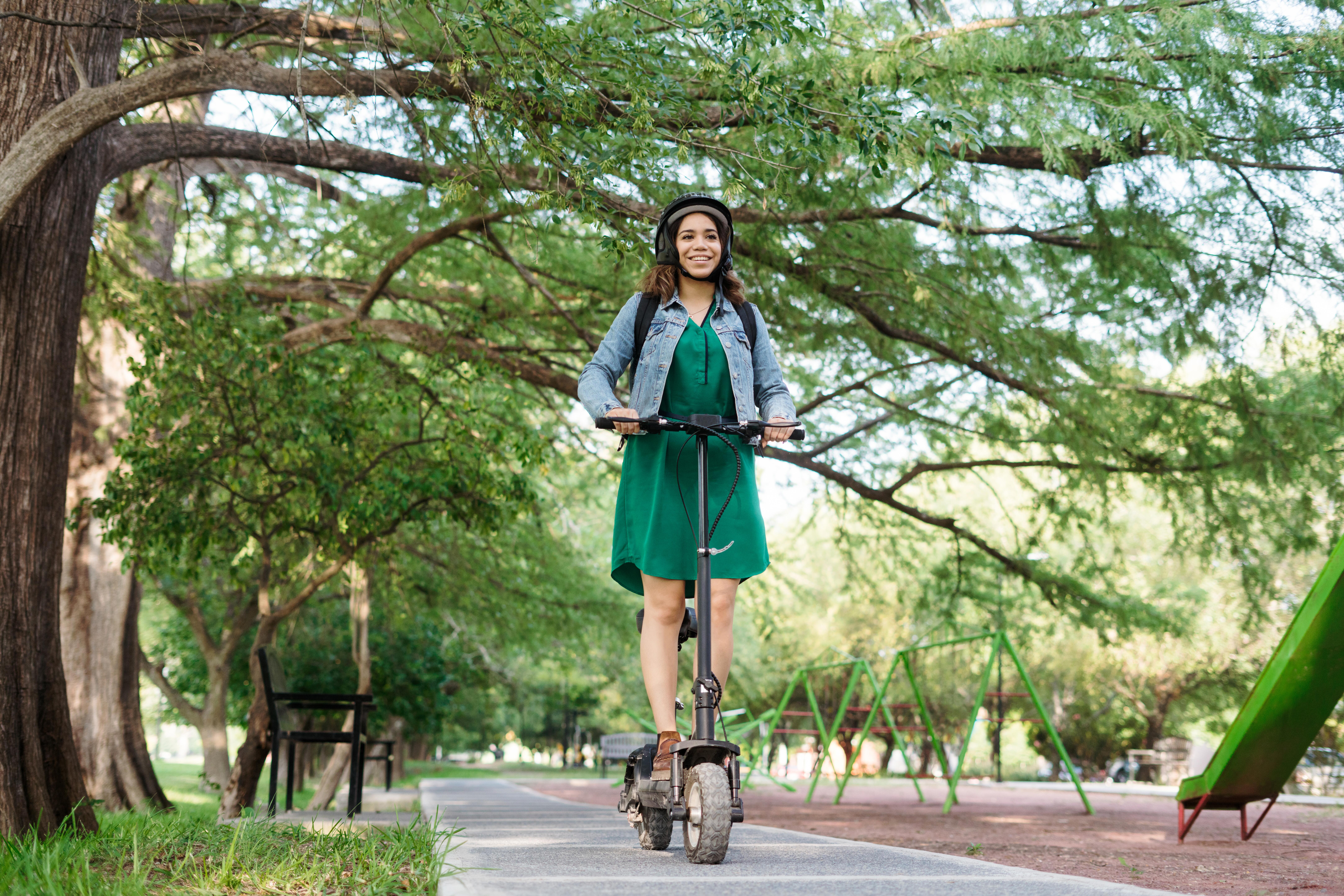 A happy young latin woman wearing a helmet and green dress, riding a motorized scooter at a park