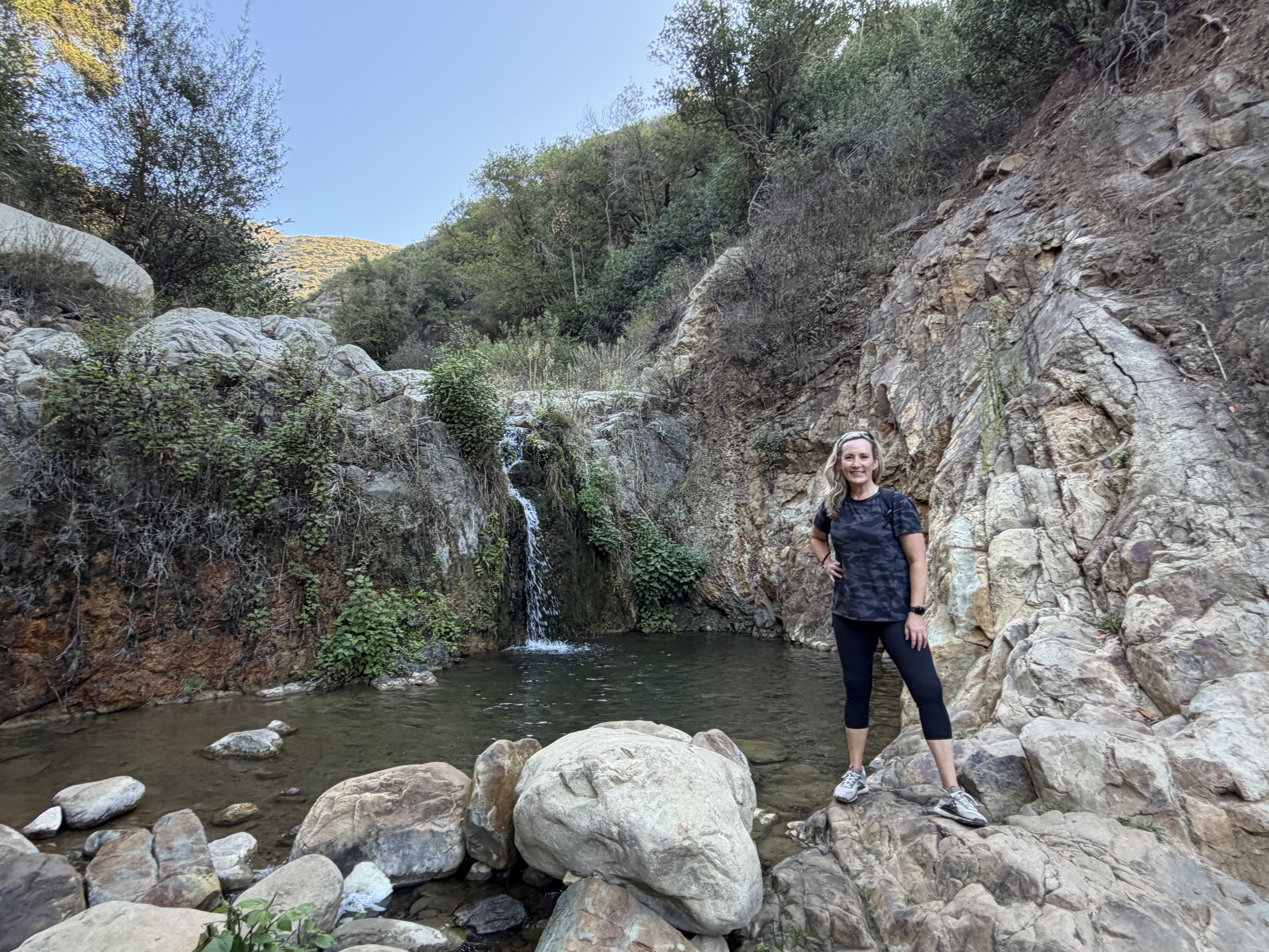 Woman with long blonde hair posed in front of small rocky waterfall.