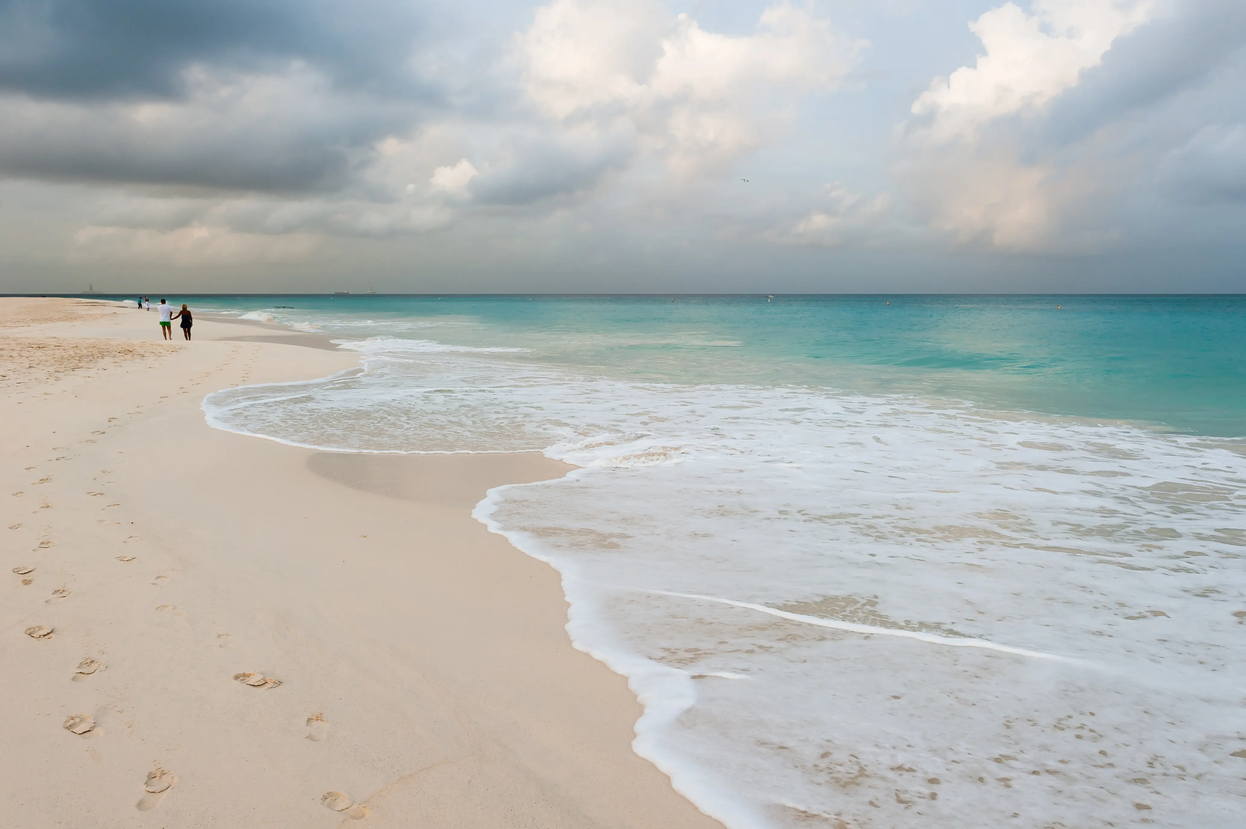 Image of two people walking in the distance on beach, with the ocean waves at the side.