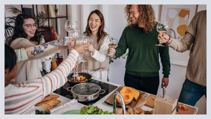 A group of friends standing around a kitchen island after cooking dinner together raise their glasses together in a toast. 