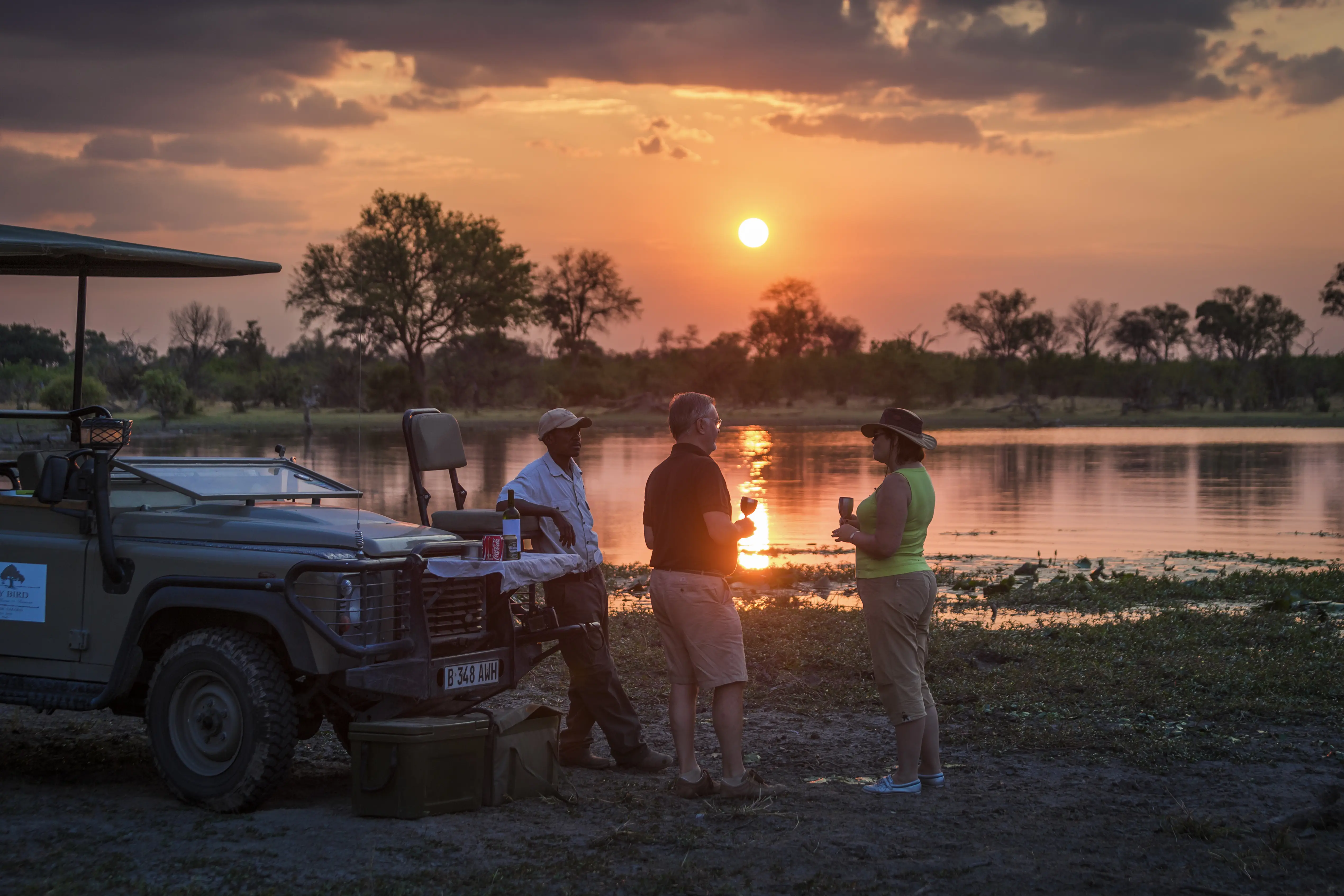 Image of tourists enjoying drinks on a safari, parked near a river.