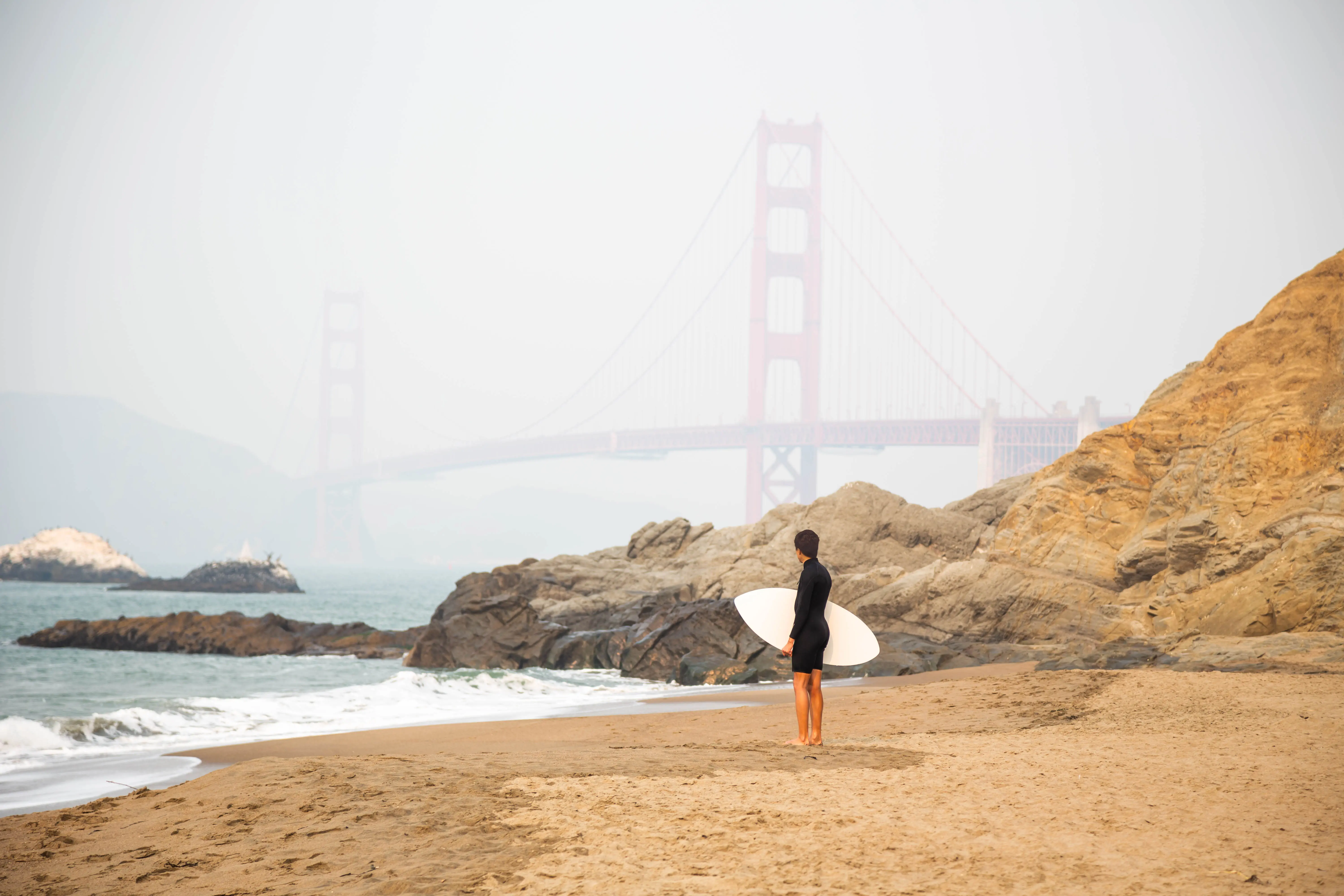 Image of a surfer standing on Baker Beach with the Golden Gate Bridge visible in the background fog.