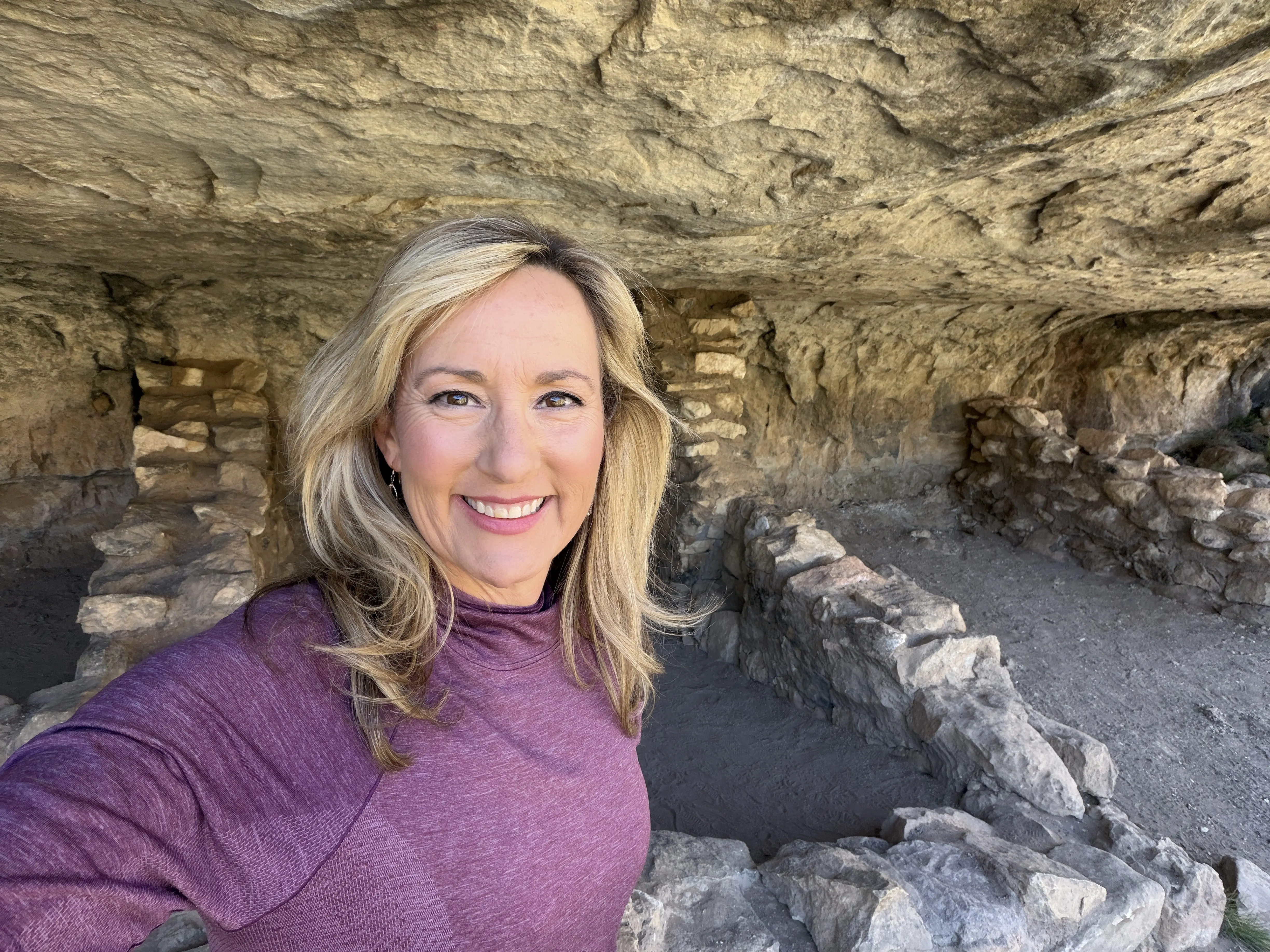 Blonde woman poses in front of ruins of ancient stone pueblo in Walnut Canyon National Monument