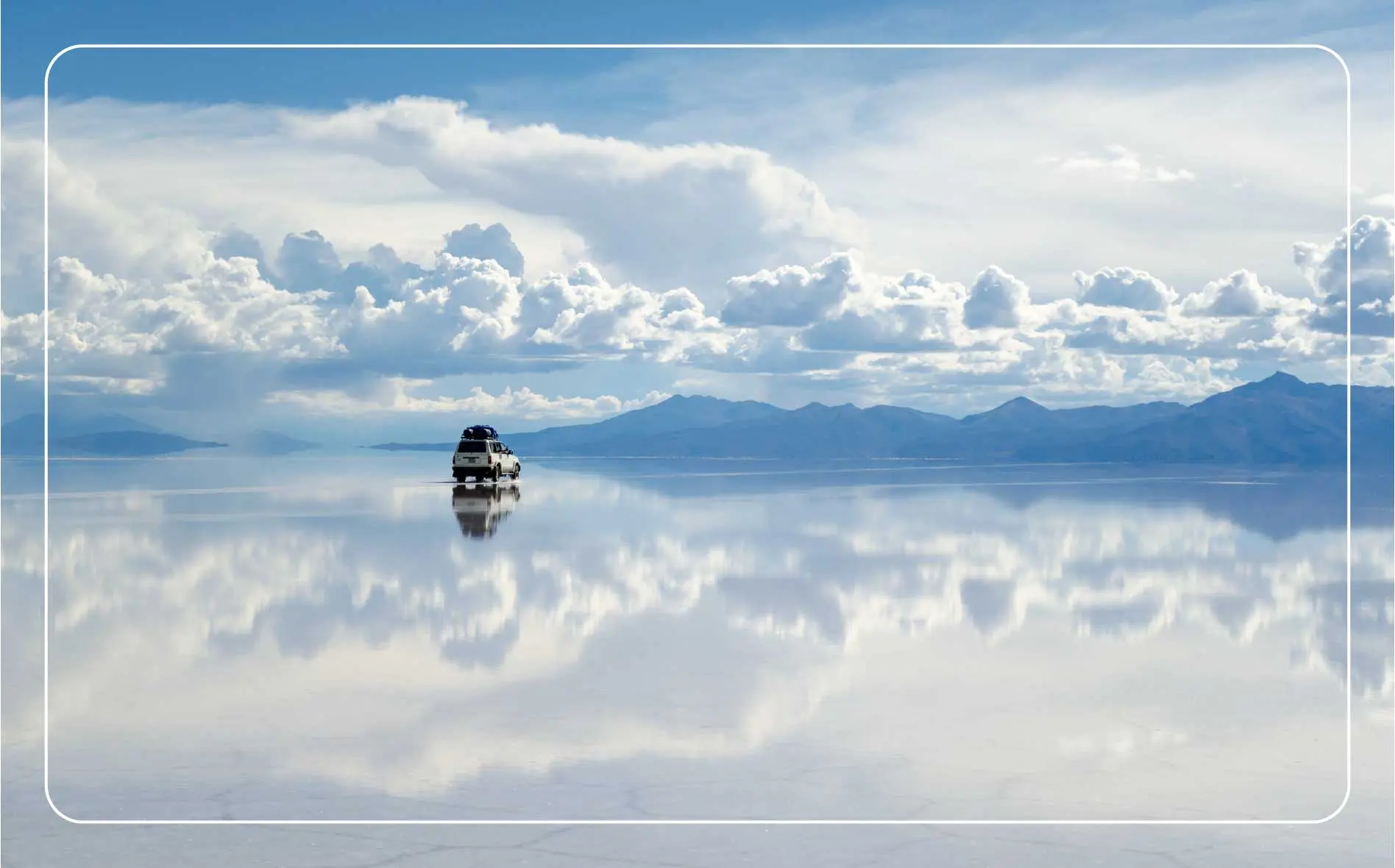 A car drives across the reflective surface of the Salar de Uyuni salt flats under a cloudy sky.