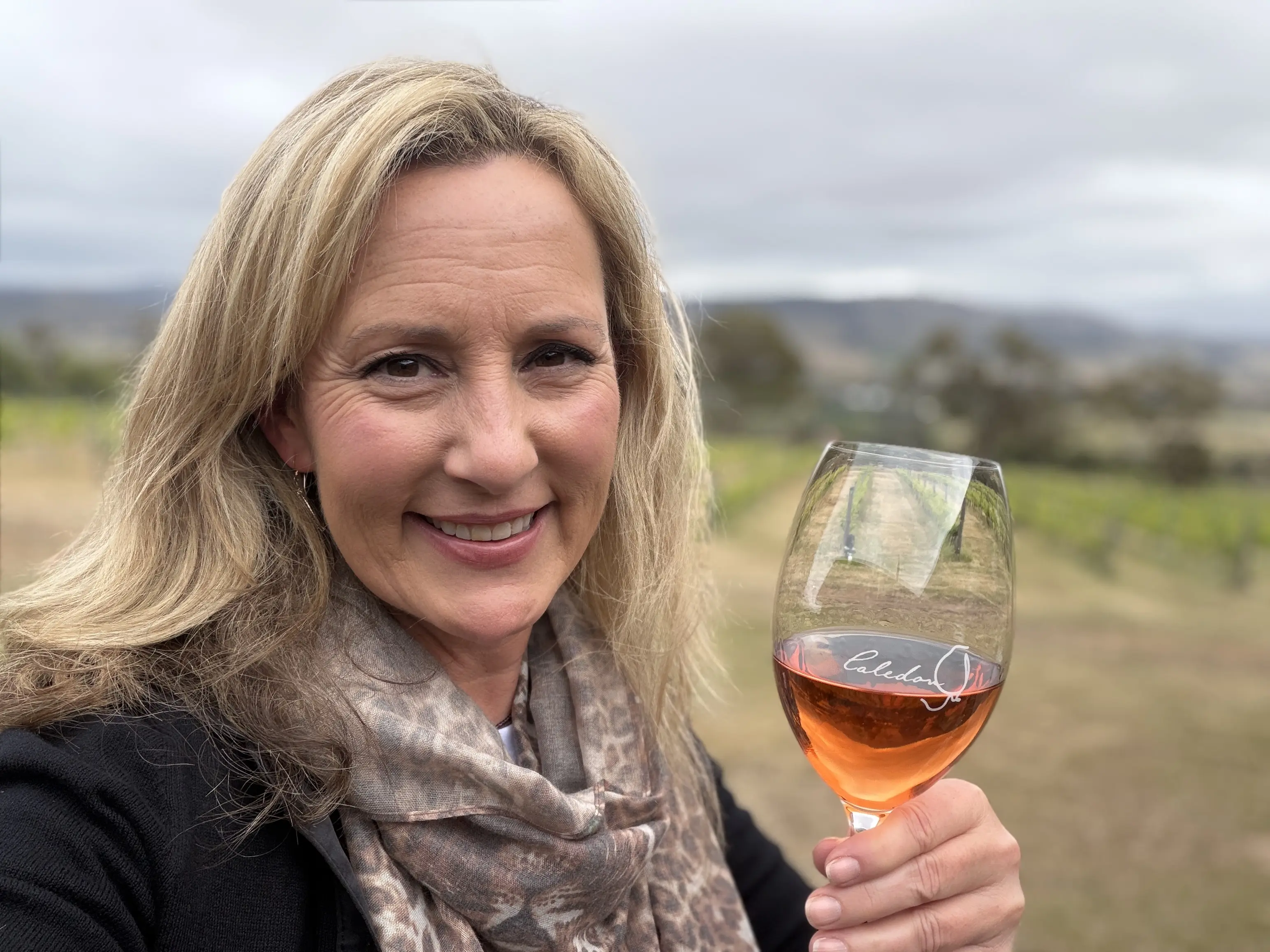 Woman with blonde hair dressed in black coat and leopard print scarf holds a glass of rosed colored wine outside in front a vineyard