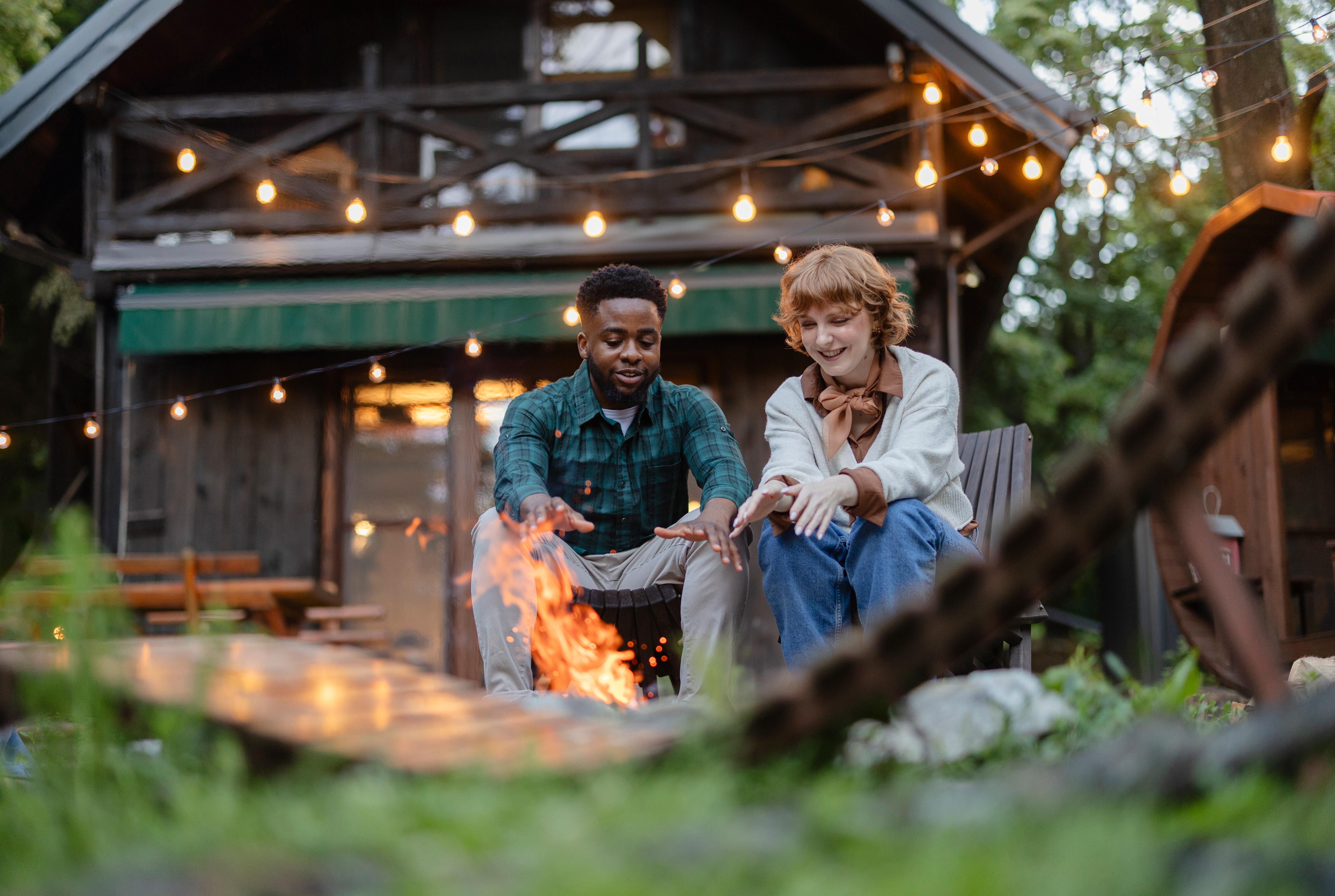 Two friends are warming their hands by a bonfire in front of a cozy wooden cabin, enjoying the warmth and ambiance during their relaxing vacation