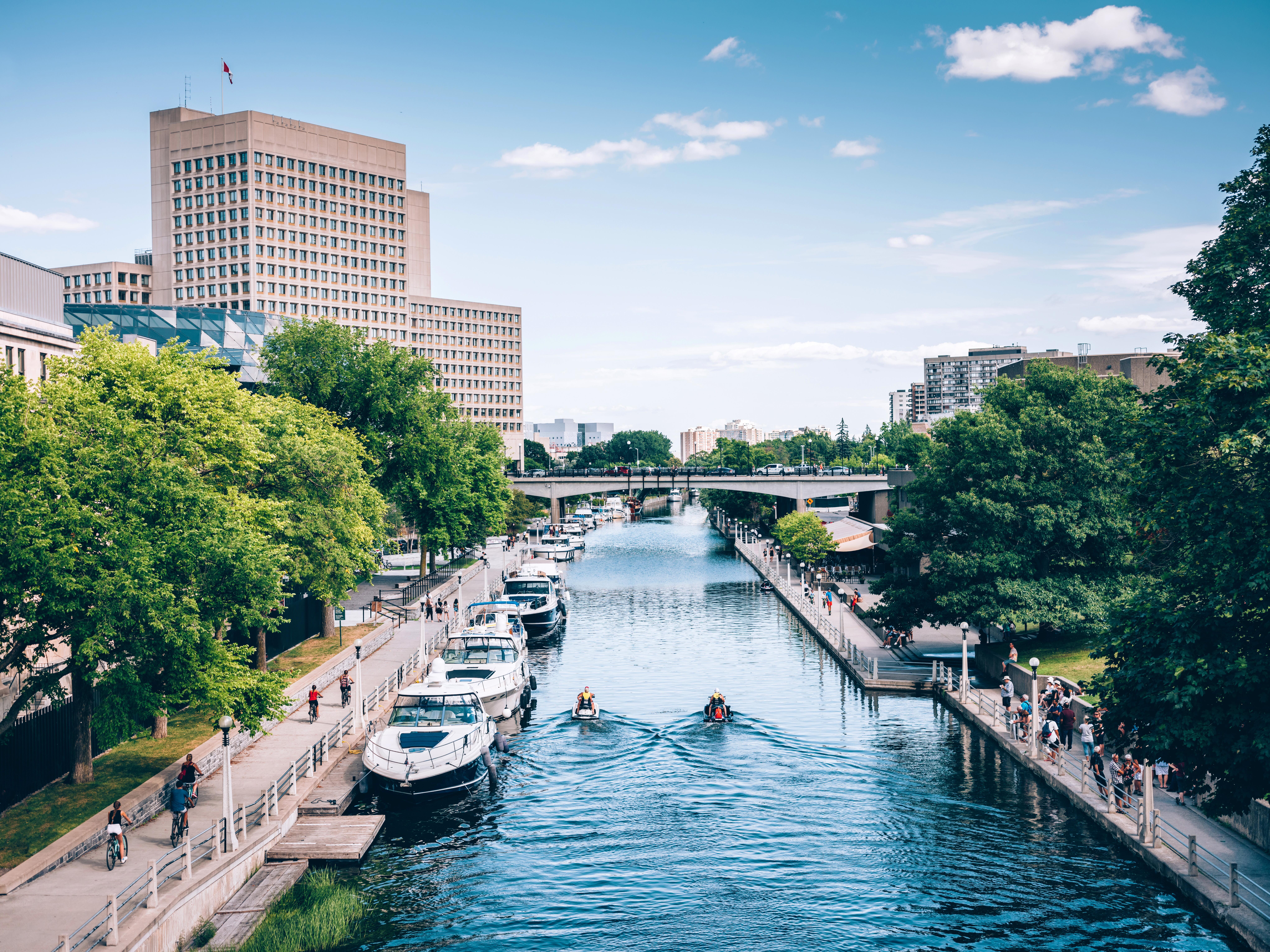 Scenic architecture of Rideau Canal and boats docked in downtown Ottawa, with people walking, biking, and wave runners on the water