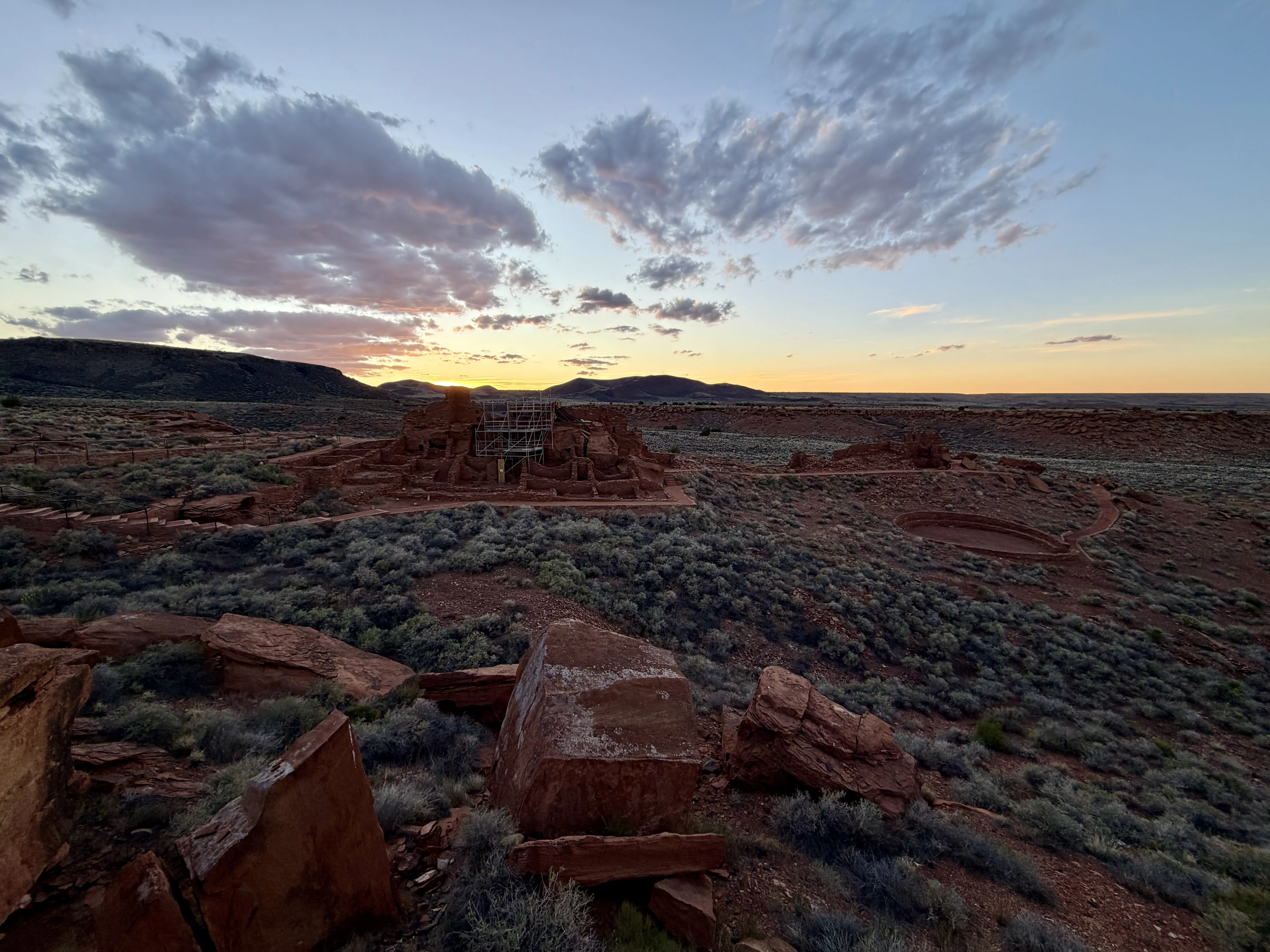 Scrubby grass fills the landscape dotted with red rocks and as the sun sets behind the mountains in the distance.
