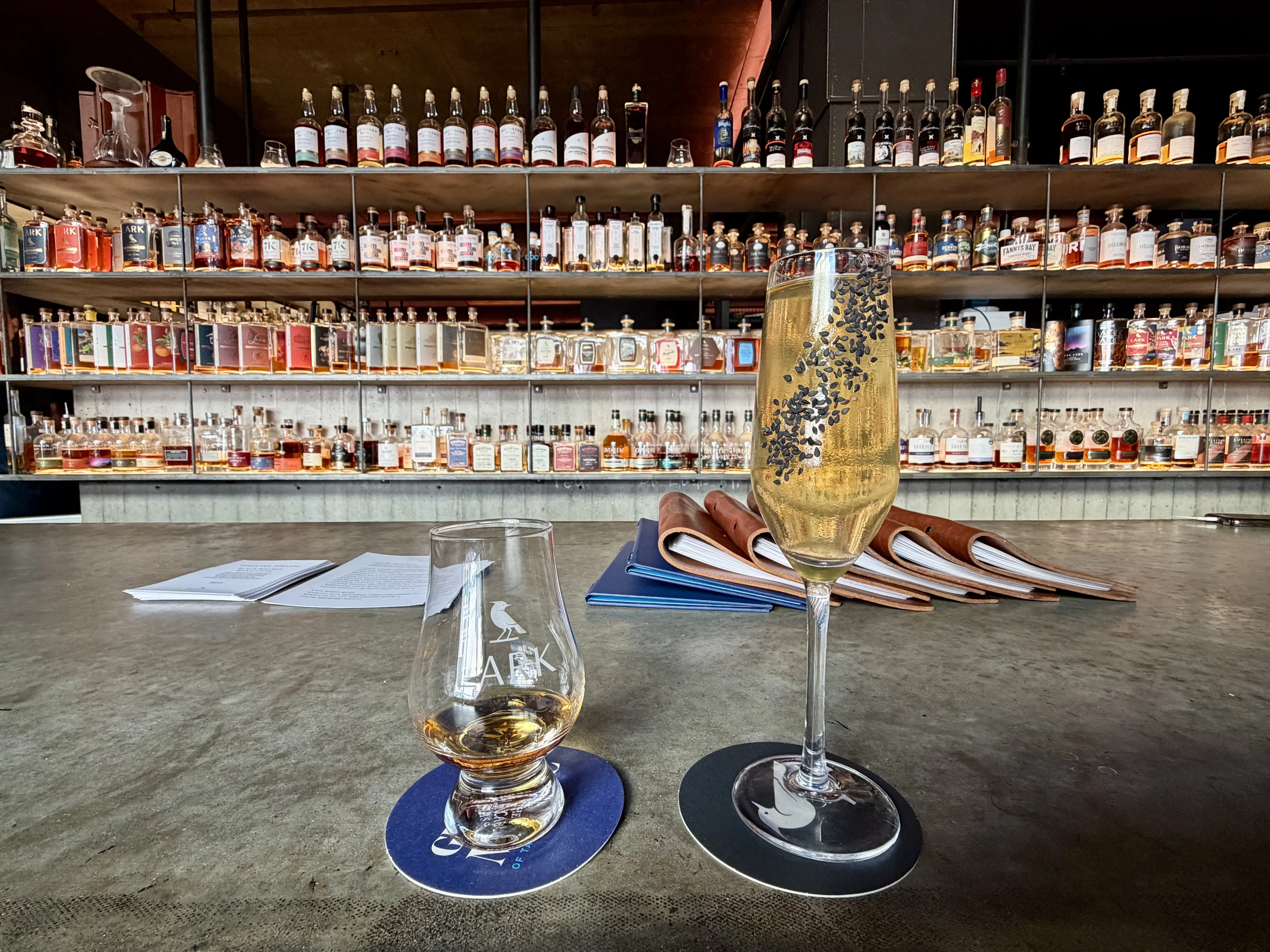 a glass of whiskey and a cocktail in a wine glass garnished with black sesame seeds atop a bar with shelves of whiskey in the background