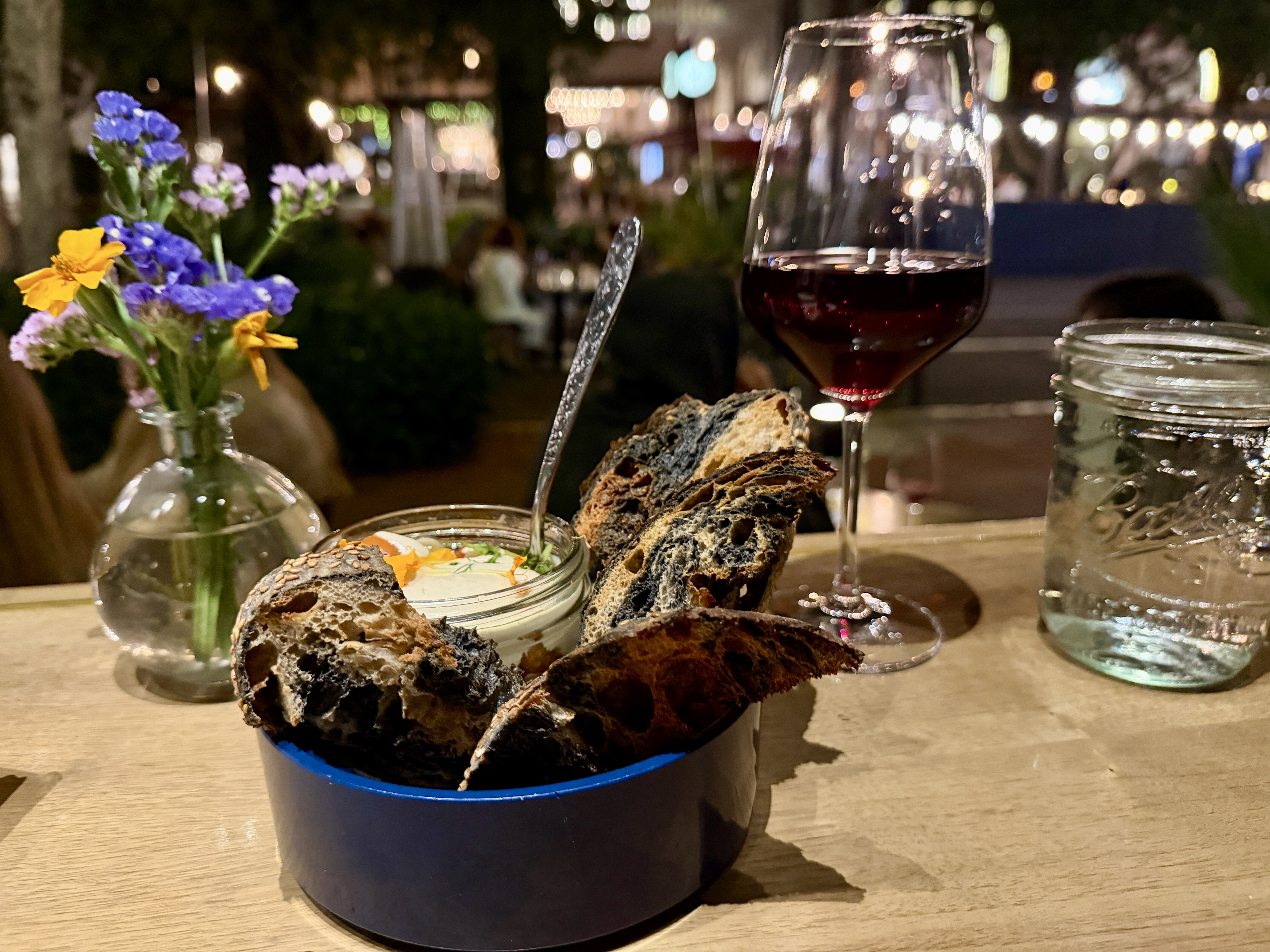 Toasted Oak Bakery sourdough with confit tomatoes on a table with red wine and a vase of flowers looking out onto a street at night.