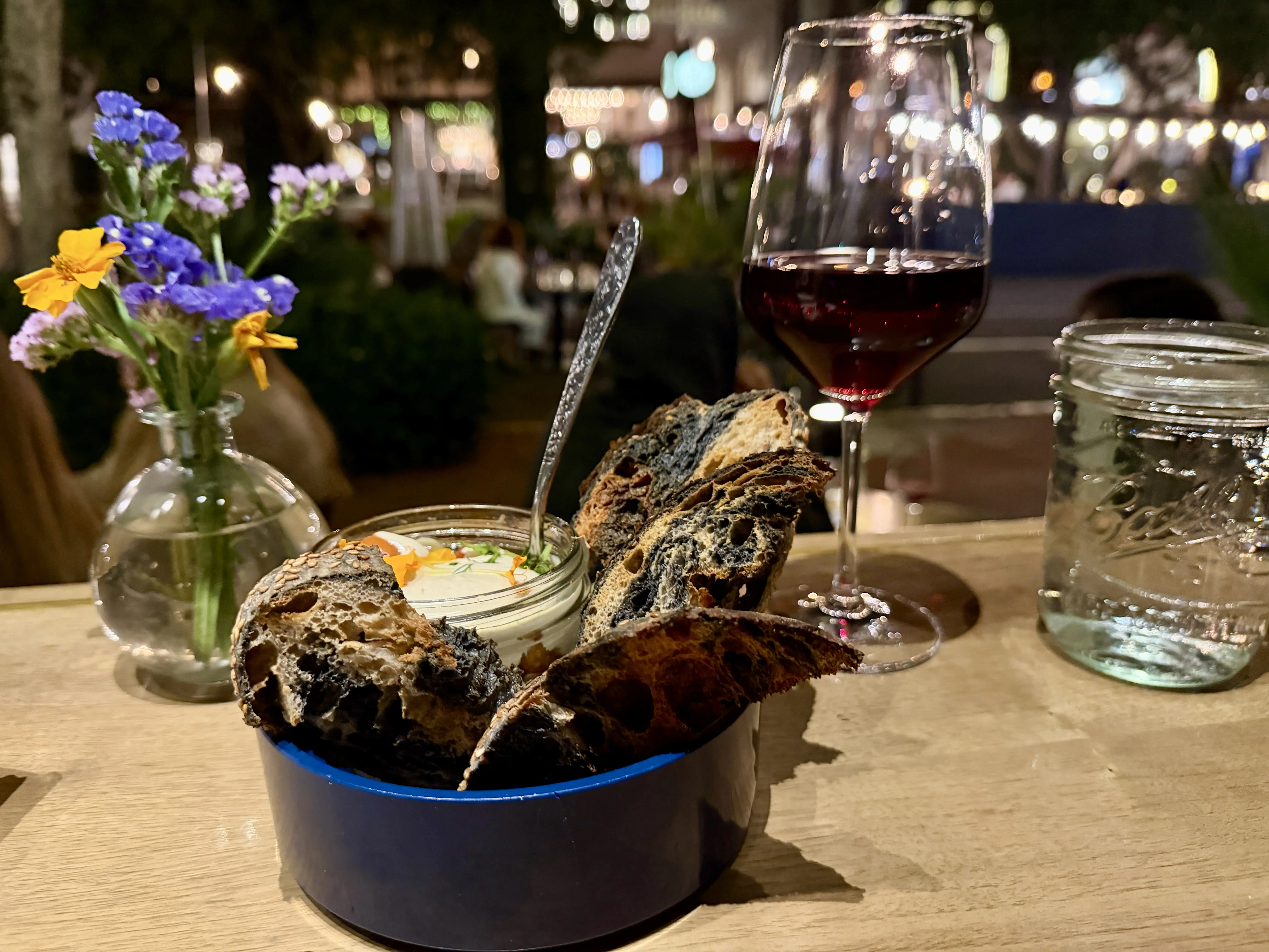 Toasted Oak Bakery sourdough with confit tomatoes on a table with red wine and a vase of flowers looking out onto a street at night.