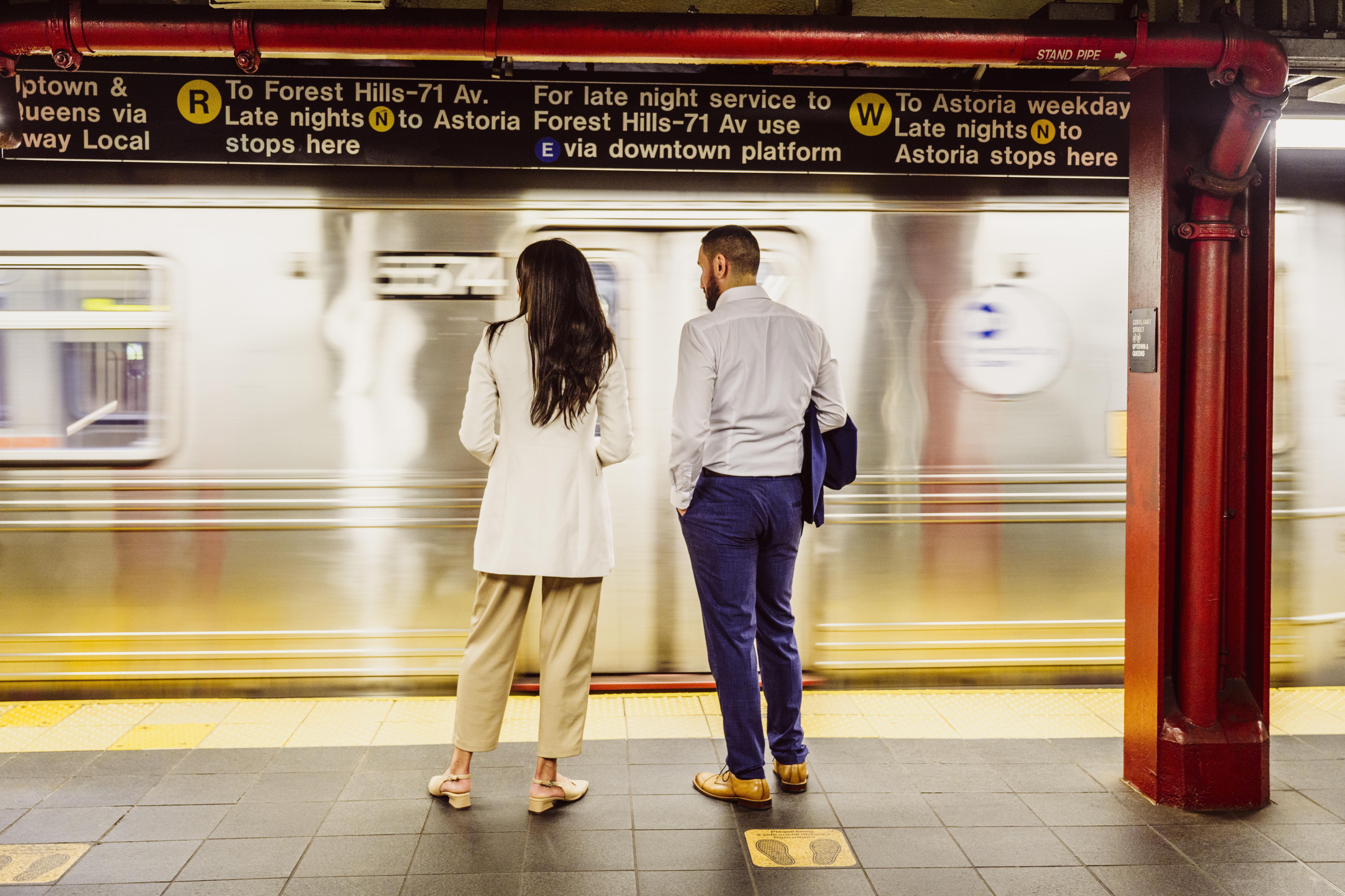 Full length rear view of young woman and mid adult man, dressed for summer, standing on platform and watching train pass, New York City
