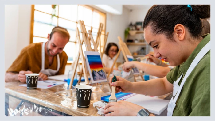 A group of people sit at a table with painting tools as they take a painting class.