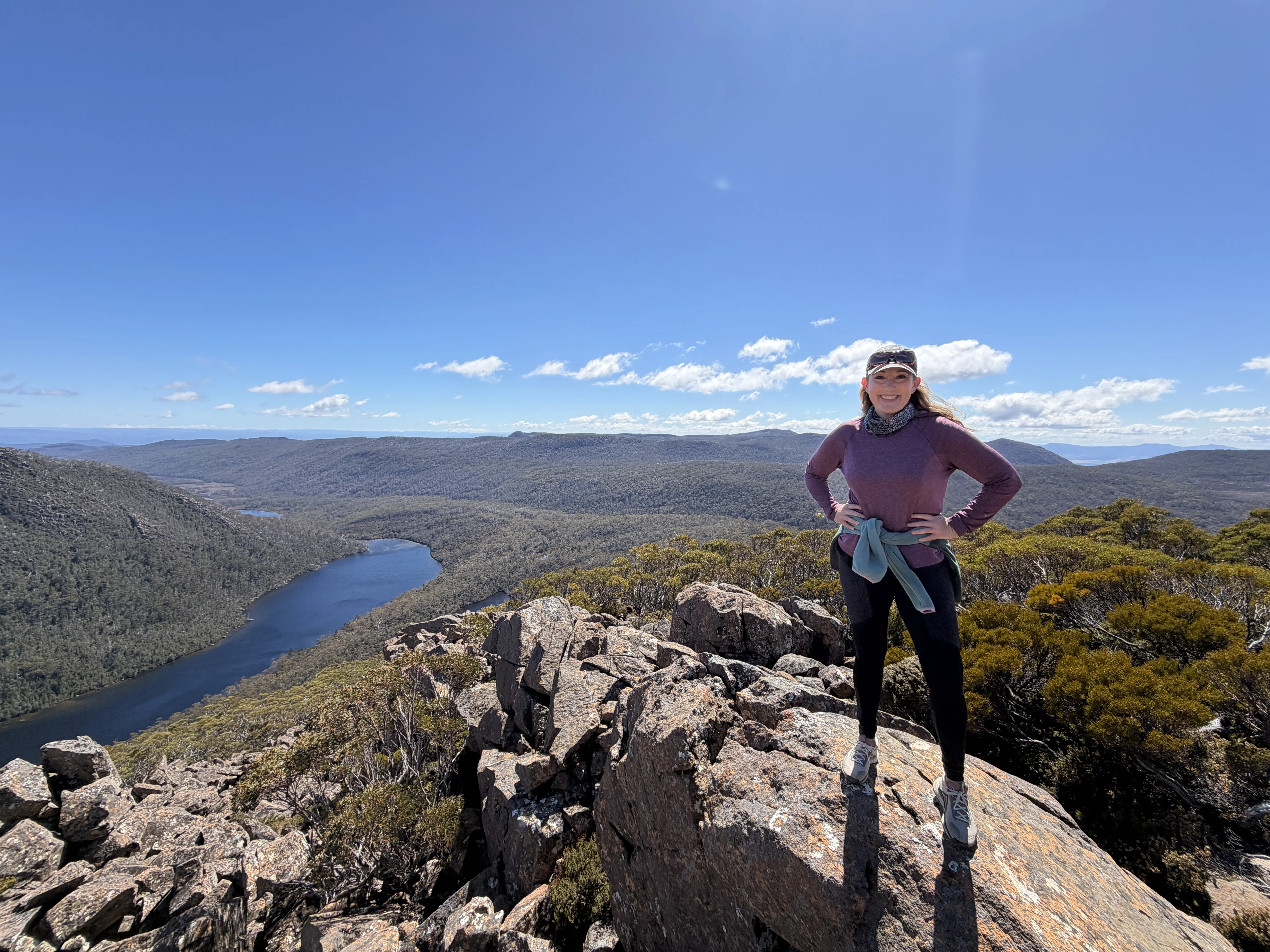 Woman with long blonde hair and dressed in hiking gear poses cliffside hands on hips. Mountains and river valley in background