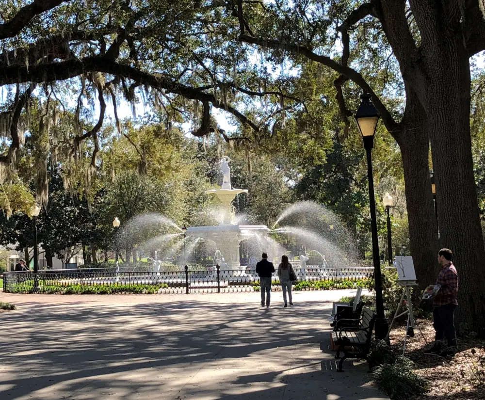 Two people walk to Savannah's Forsyth Park fountain while artist paints landscape.