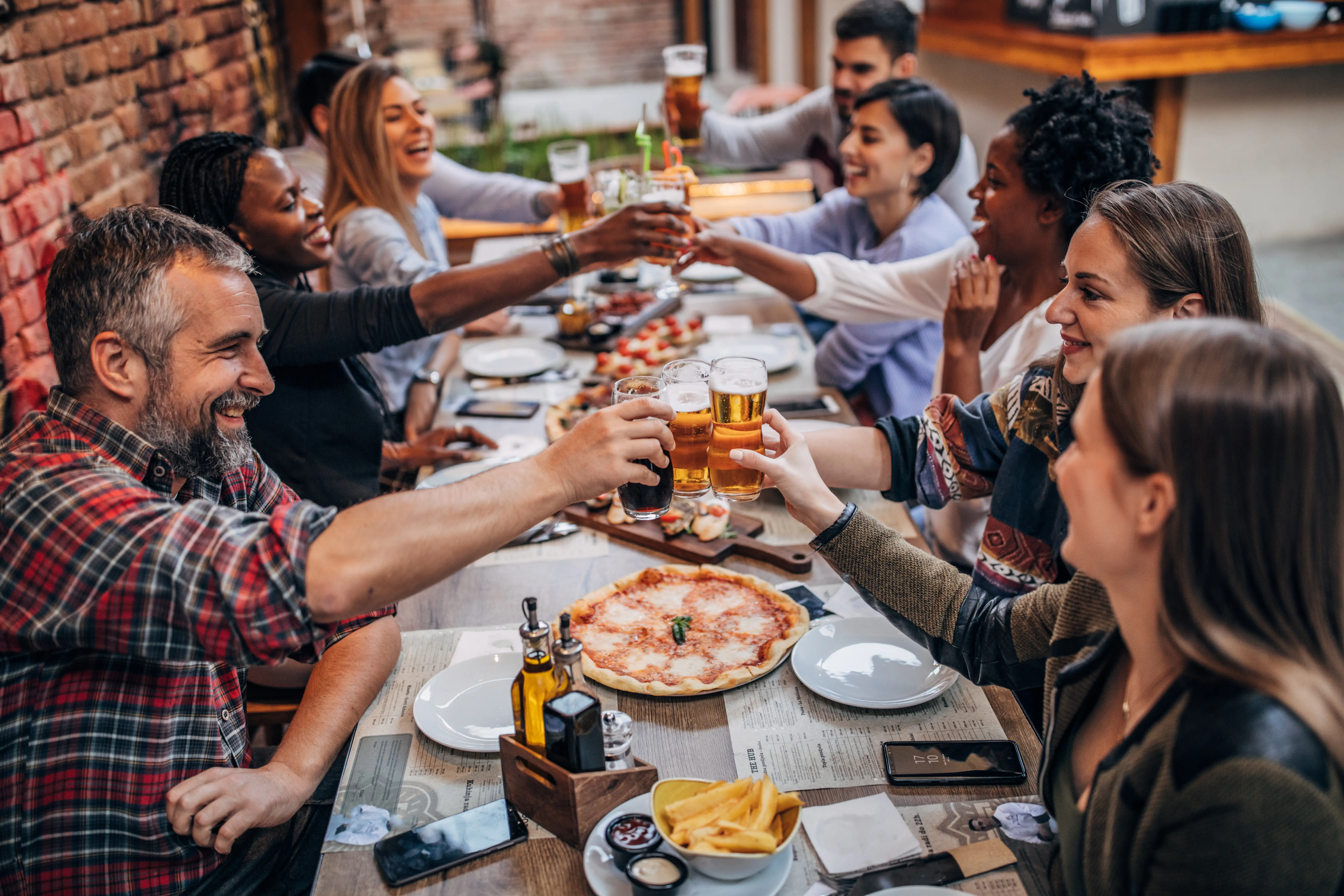Ethnic group of friends dining in a rustic pub, raising their glasses to express good wishes, with pizza, flatbread and ale on a wooden table