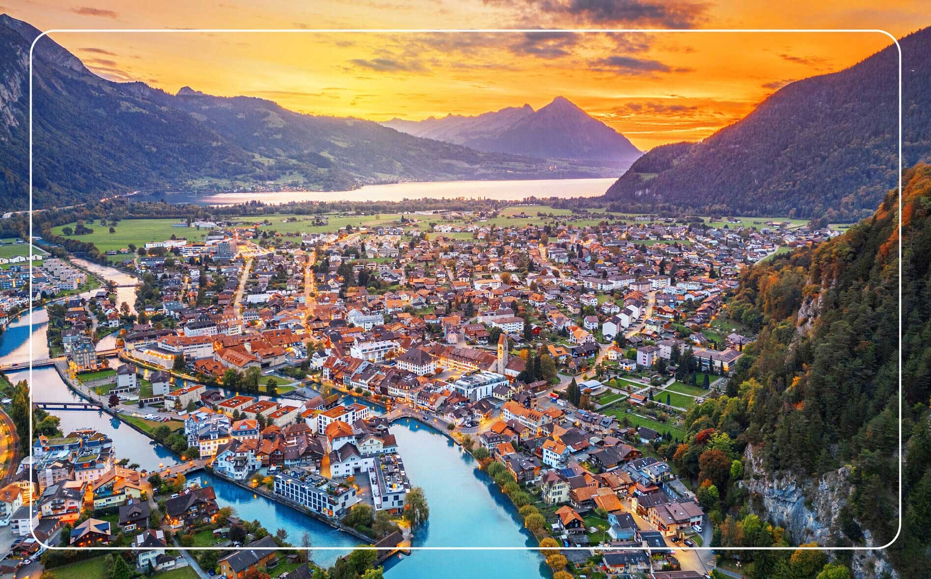 Aerial view of Interlaken at golden hour, showcasing orange-roofed buildings nestled between a turquoise alpine lake and mountains in the background.