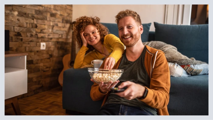 A couple sits in their living room eating a bowl of popcorn before an at-home movie night.