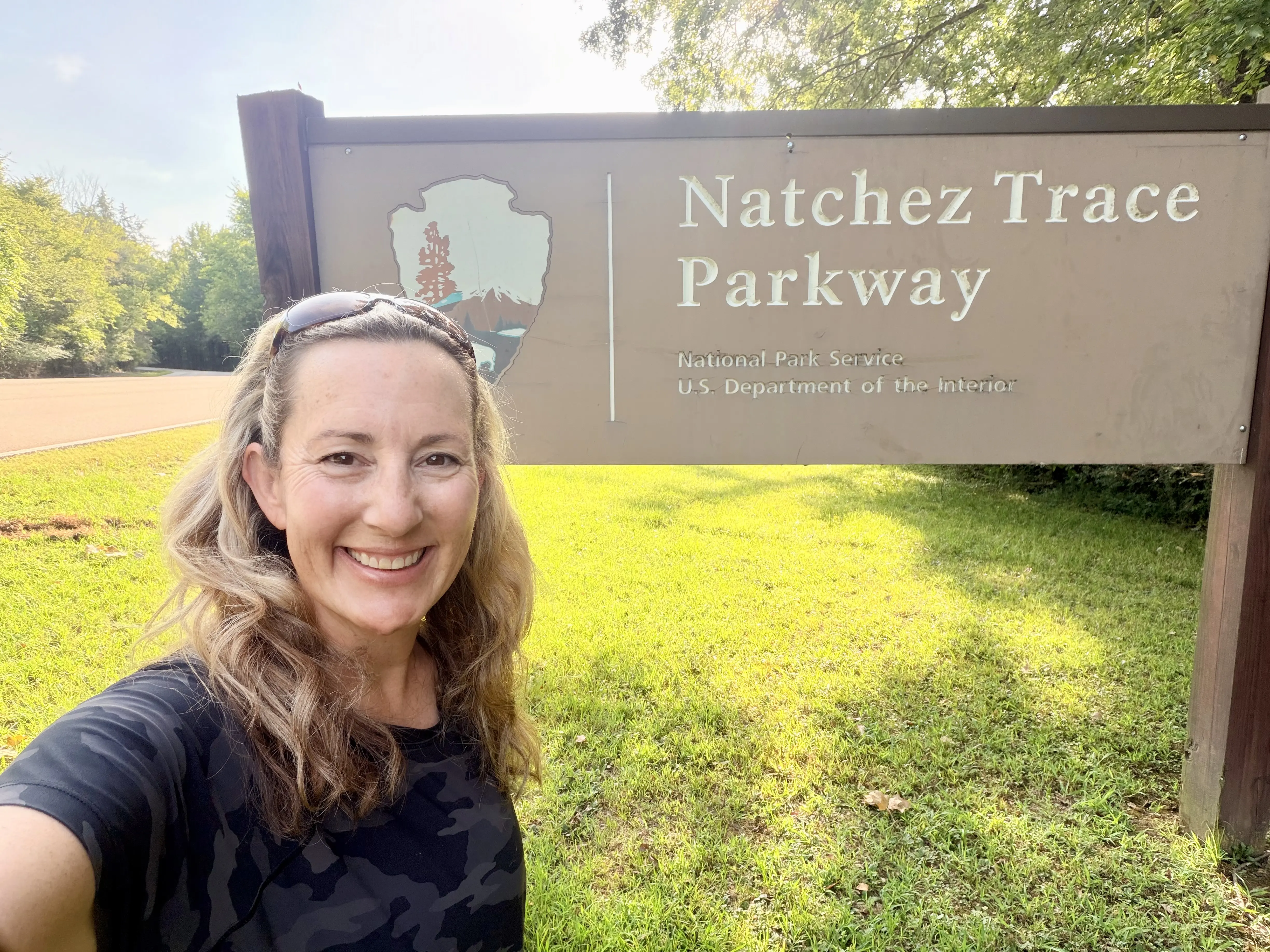 Woman with long blond hair in black t-shirt stand atop grass in front of a sign with a National Park Service badge and in large letters says, "Natchez Trace Parkway."