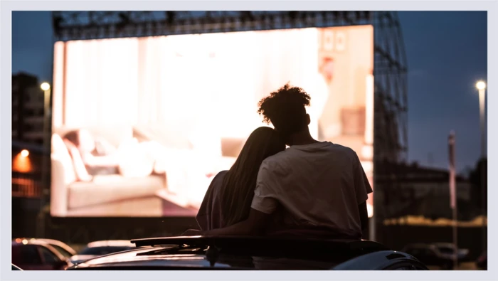 A couple sits on top of their car watching a drive-in movie. 