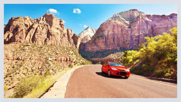 A car drives along an open road with mountains of a national park shown in the background. 