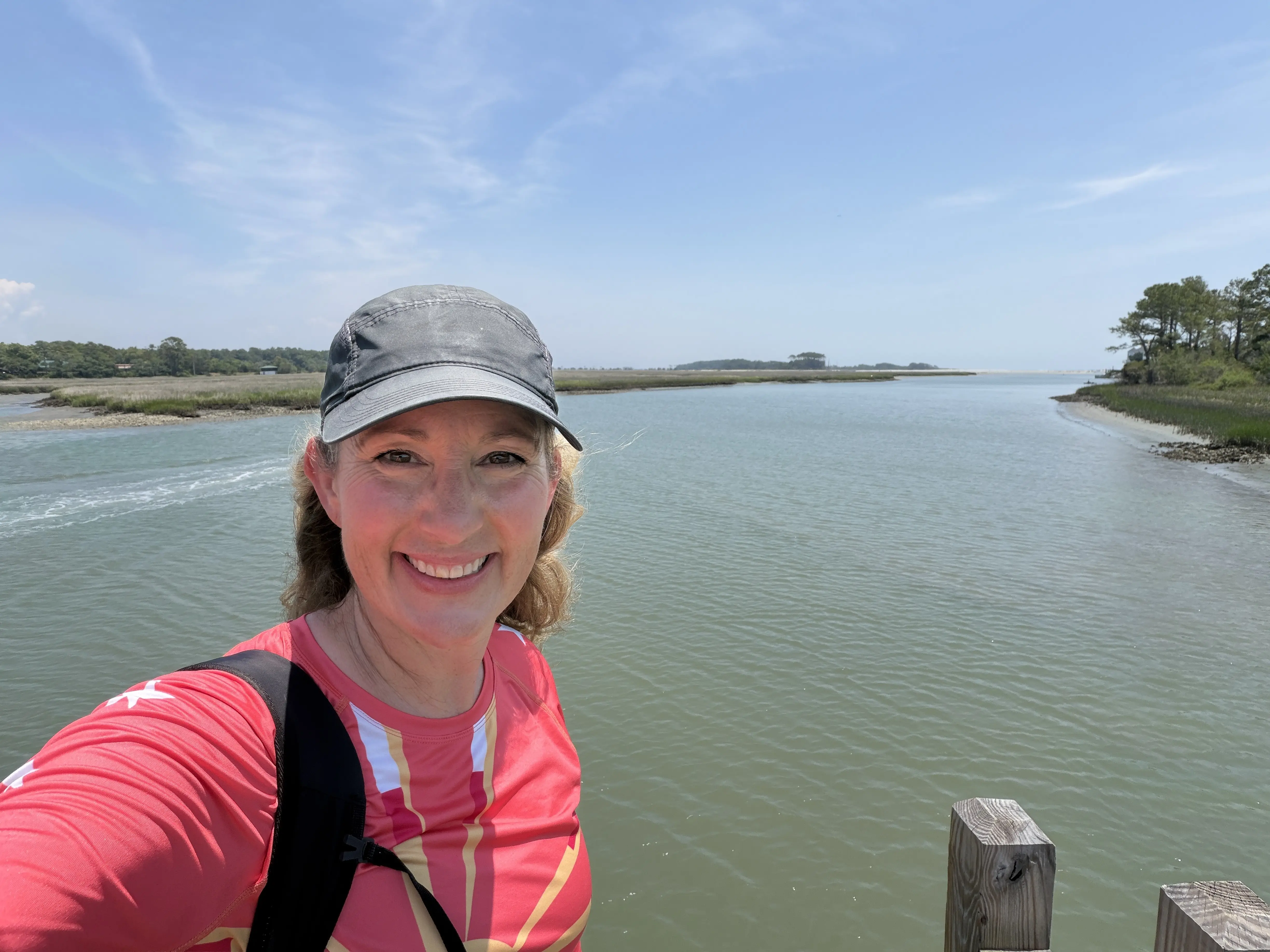 Blonde woman in black cap and pink long sleave shirt standing on a small pier in a marsh