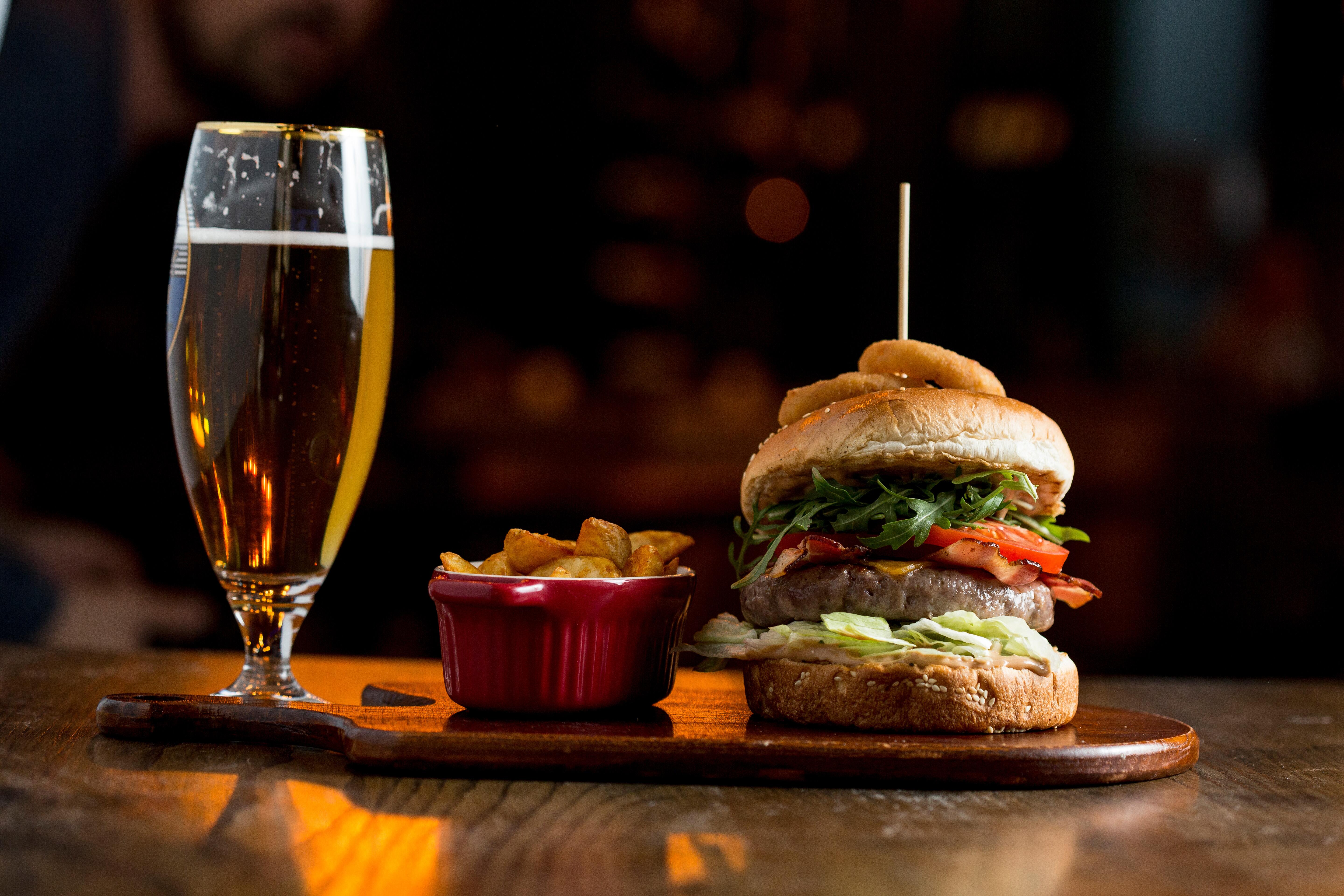 Warm light view of a beef burger in homemade pizza dough bun with BBQ sauce, melted cheddar cheese, bacon, tomato, lettuce topped with onion rings, spicy fries and ale on the table