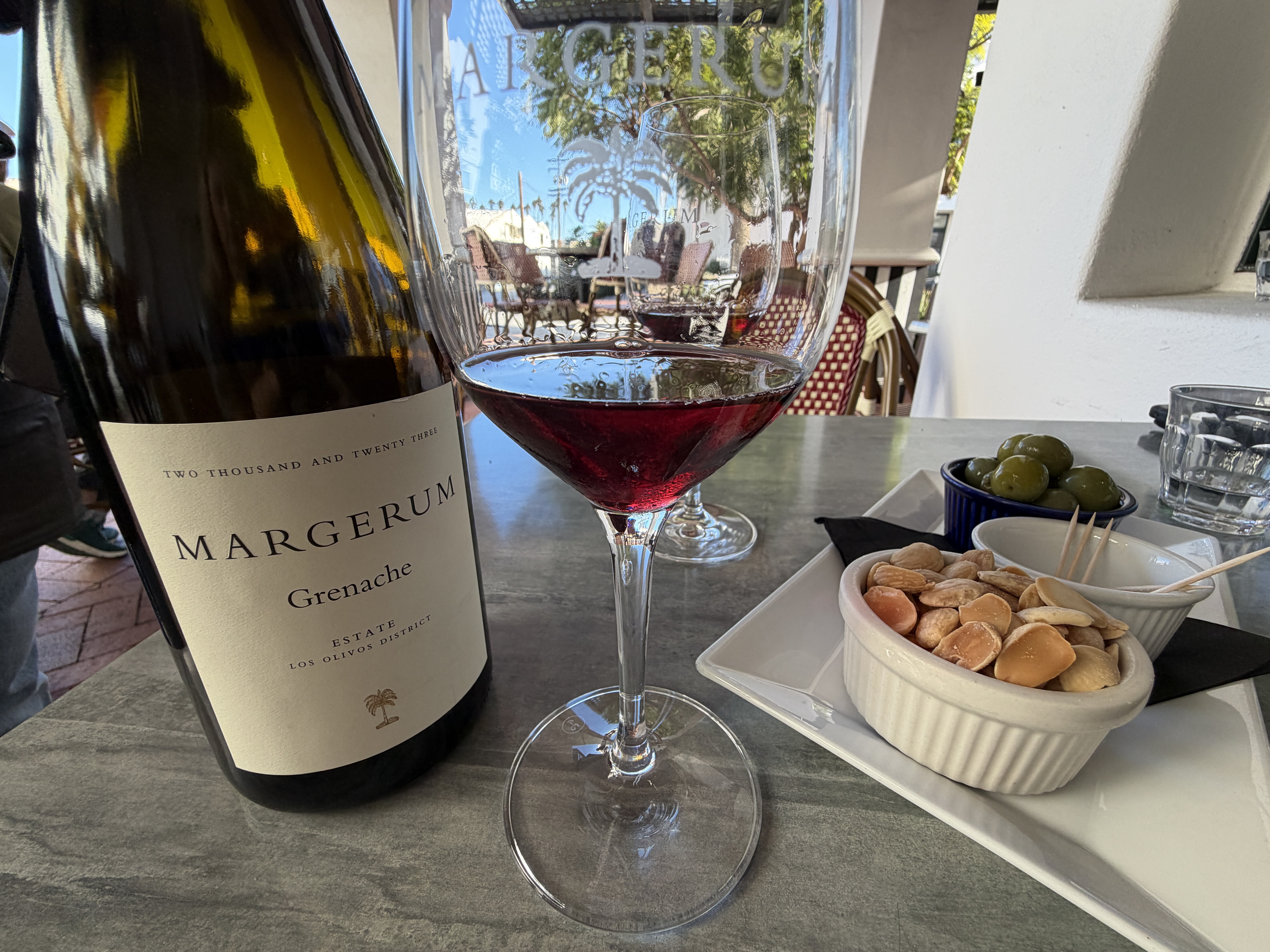 A glass of red wine and a bottle of Margerum Grenache wine atop a patio table with sunny blue sky and palm trees in background