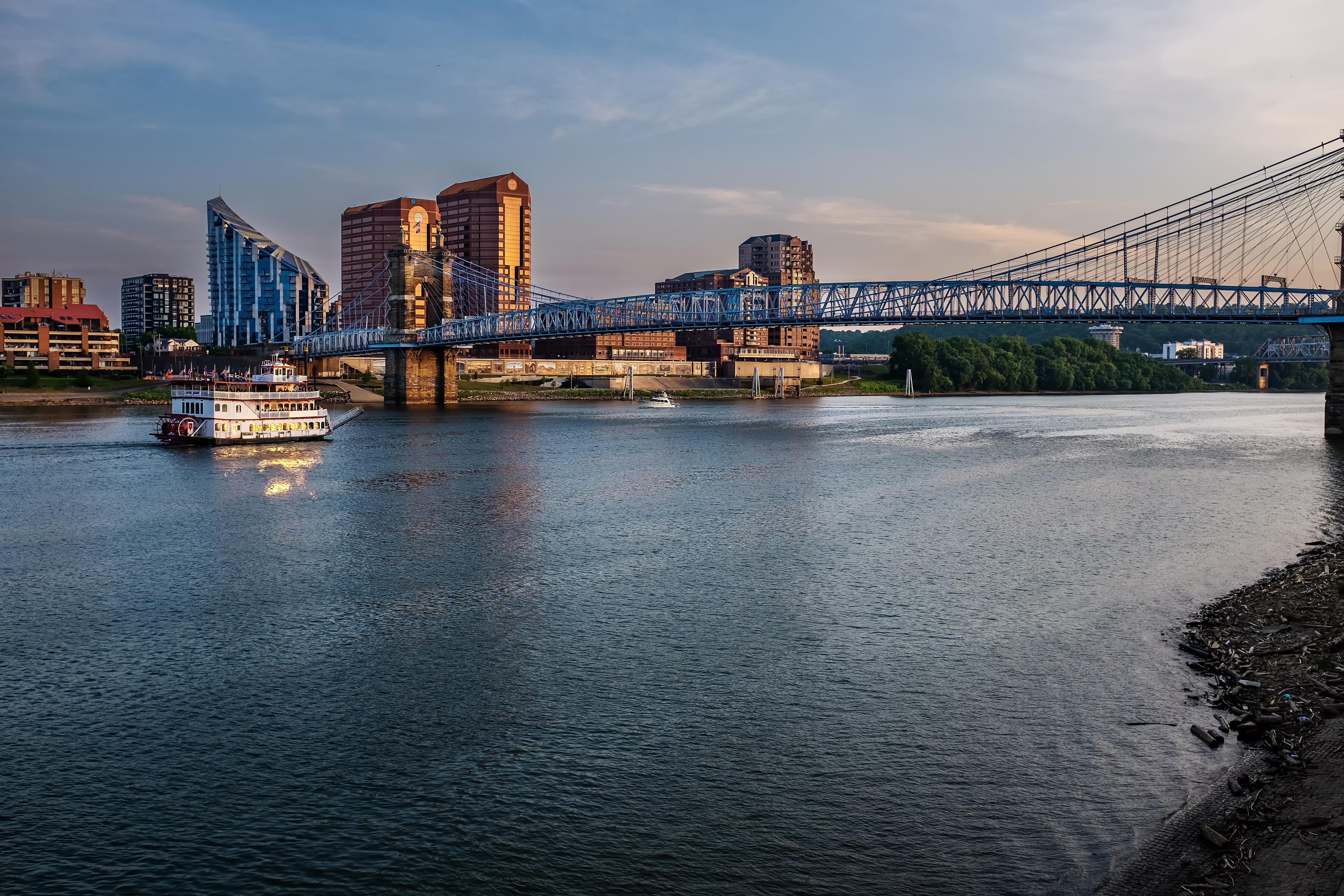 Image of a paddle boat river cruise sailing the Ohio River as it passes under old suspension bridge between Cincinnati, Ohio and Covington, Kentucky.