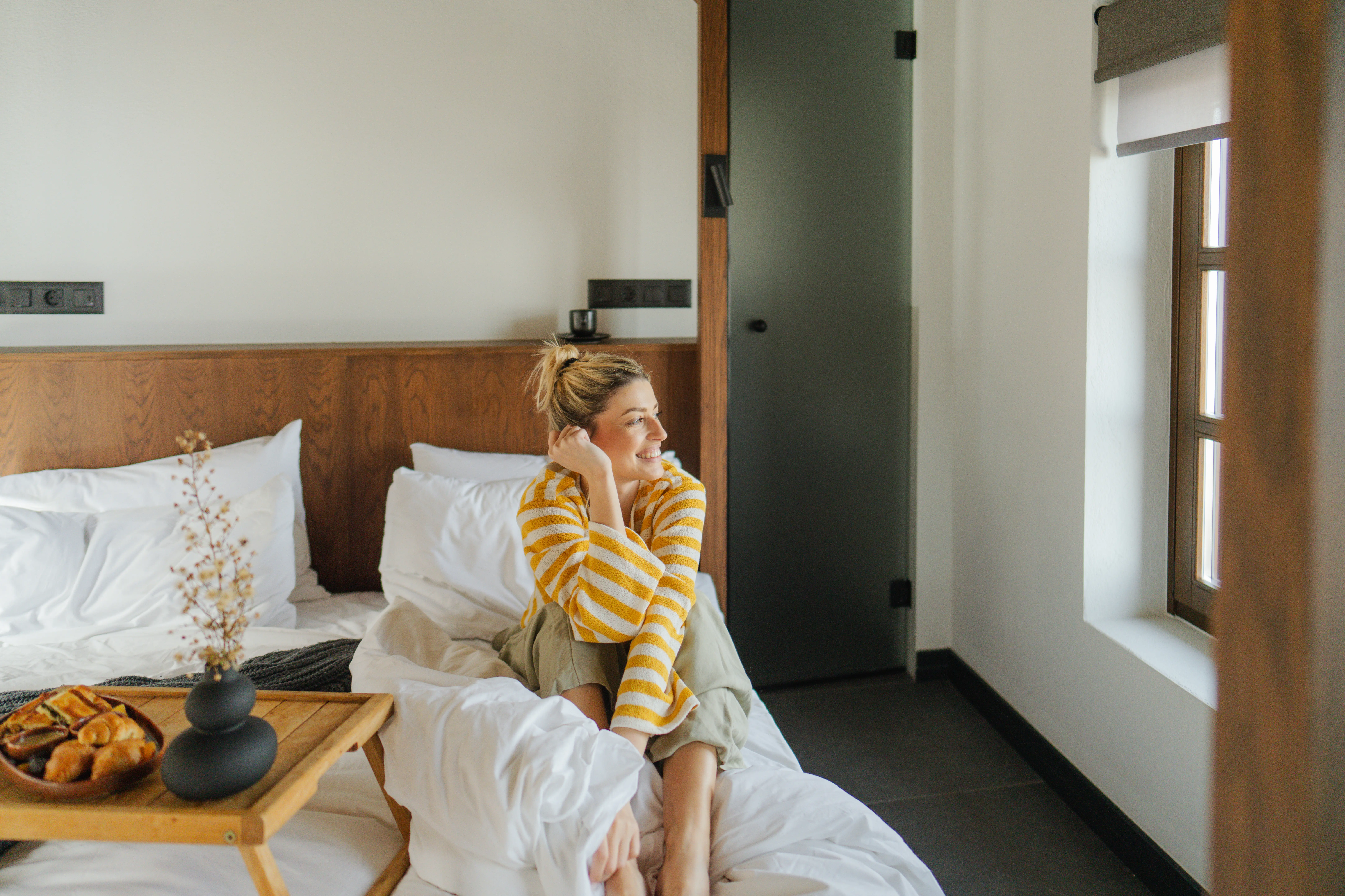 Photo of a young woman waking up, stretching and welcoming morning sun while still in bed, while breakfast platter is by her side.