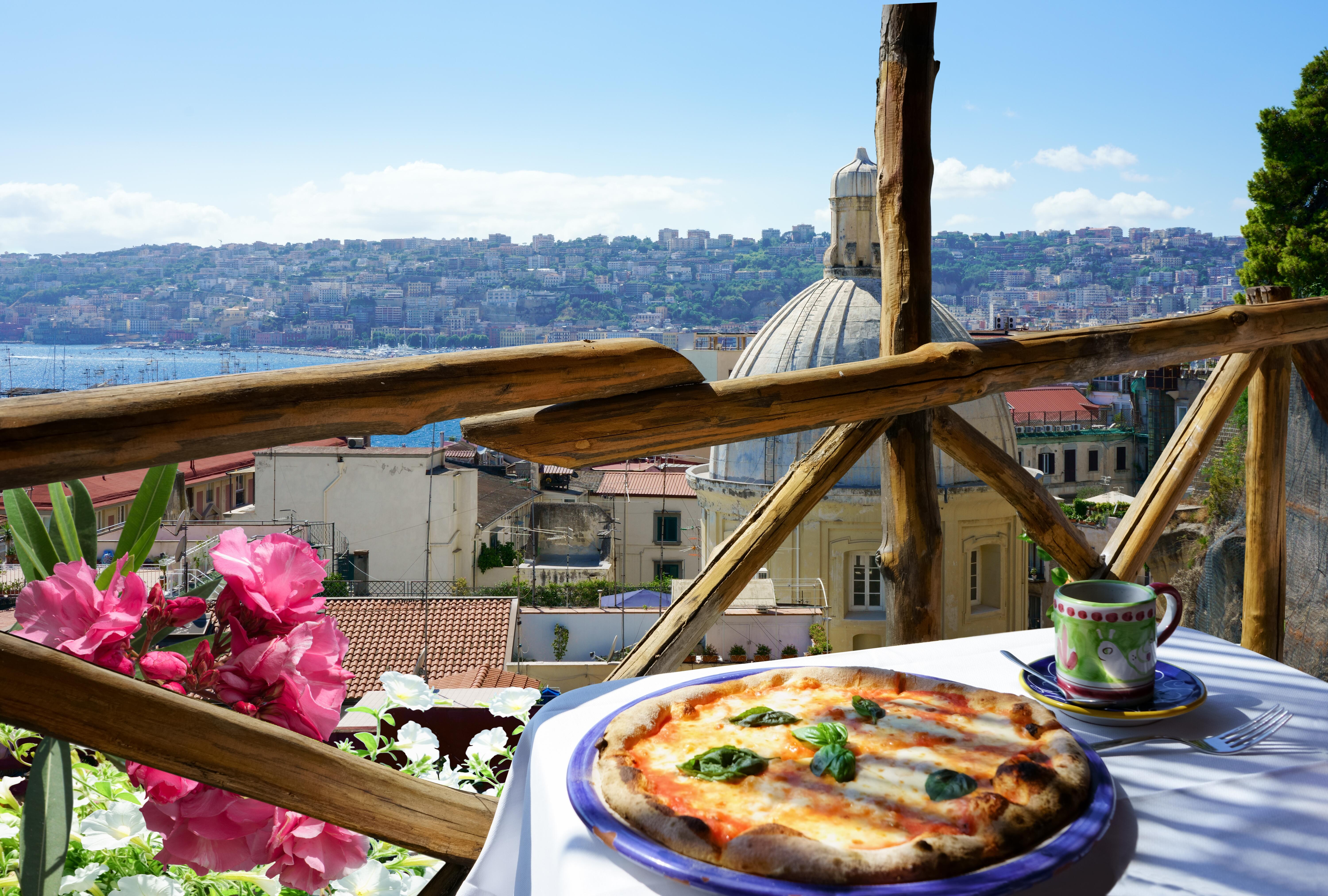 Sunny day view of pizza on a table next to pink and white flowers overlooking Naples city, Italy