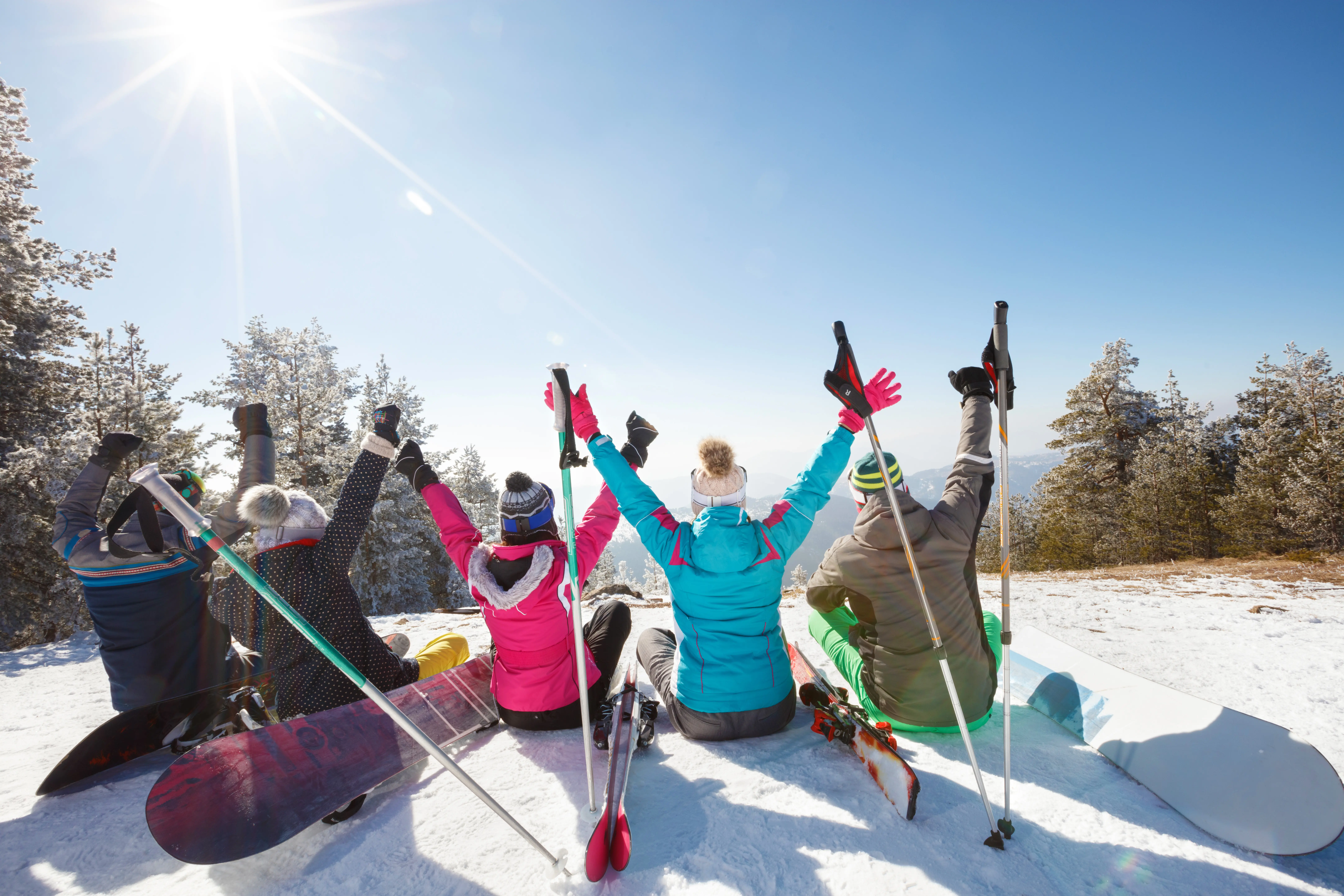 Back view of skiers sitting on a snowy hilltop with hands up and resting, on a sunny day with blue sky
