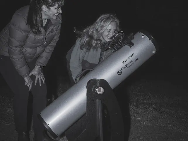 Black and white photo of woman with long hair peering through a telescope pointed at the night sky