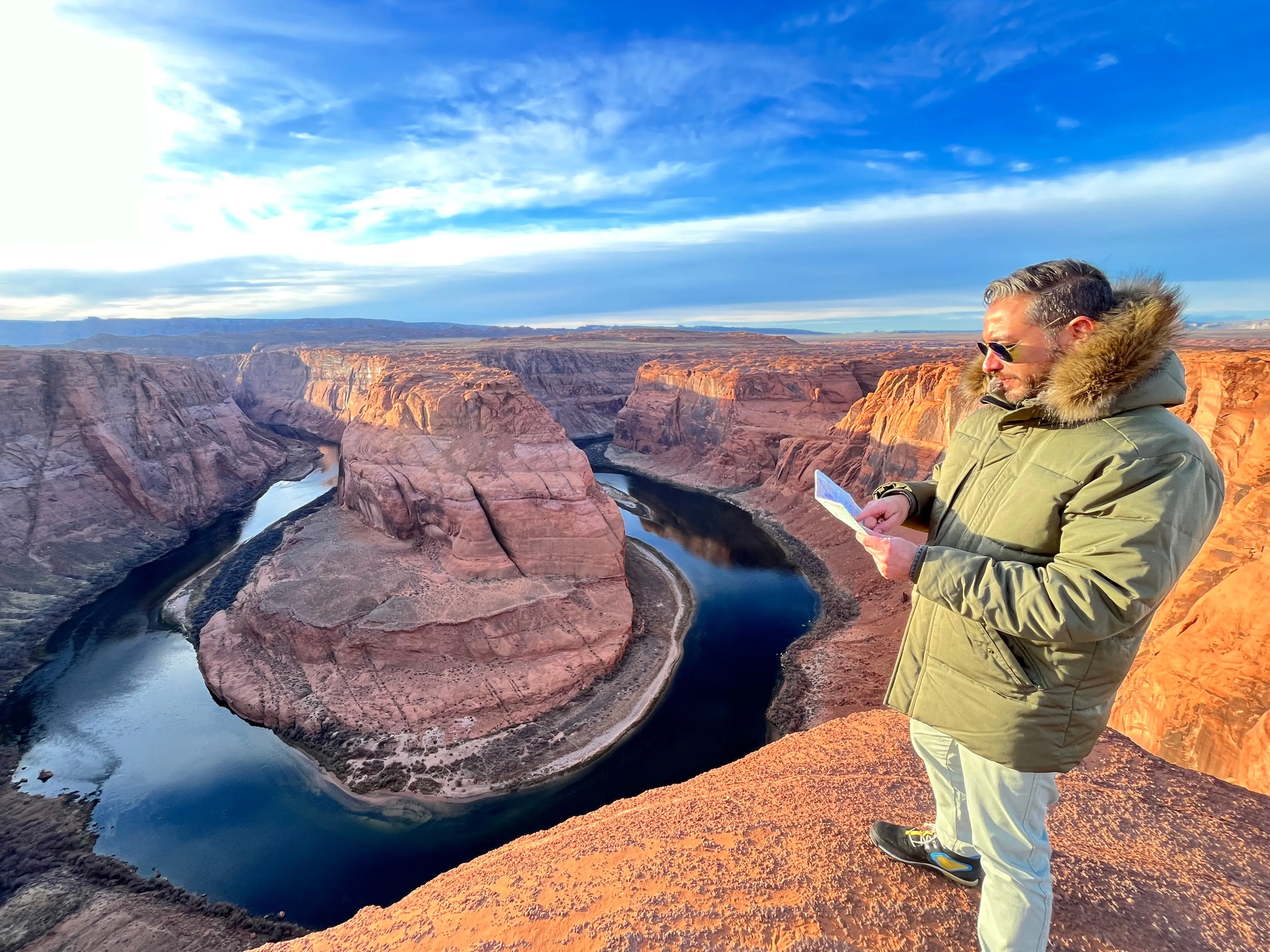 Young male traveler in green jacket and black boots during winter, on top of Horseshoe Bend, while looking the horizon checking on a map the landscape of Glen / Grand Canyon National Park in Arizona, USA