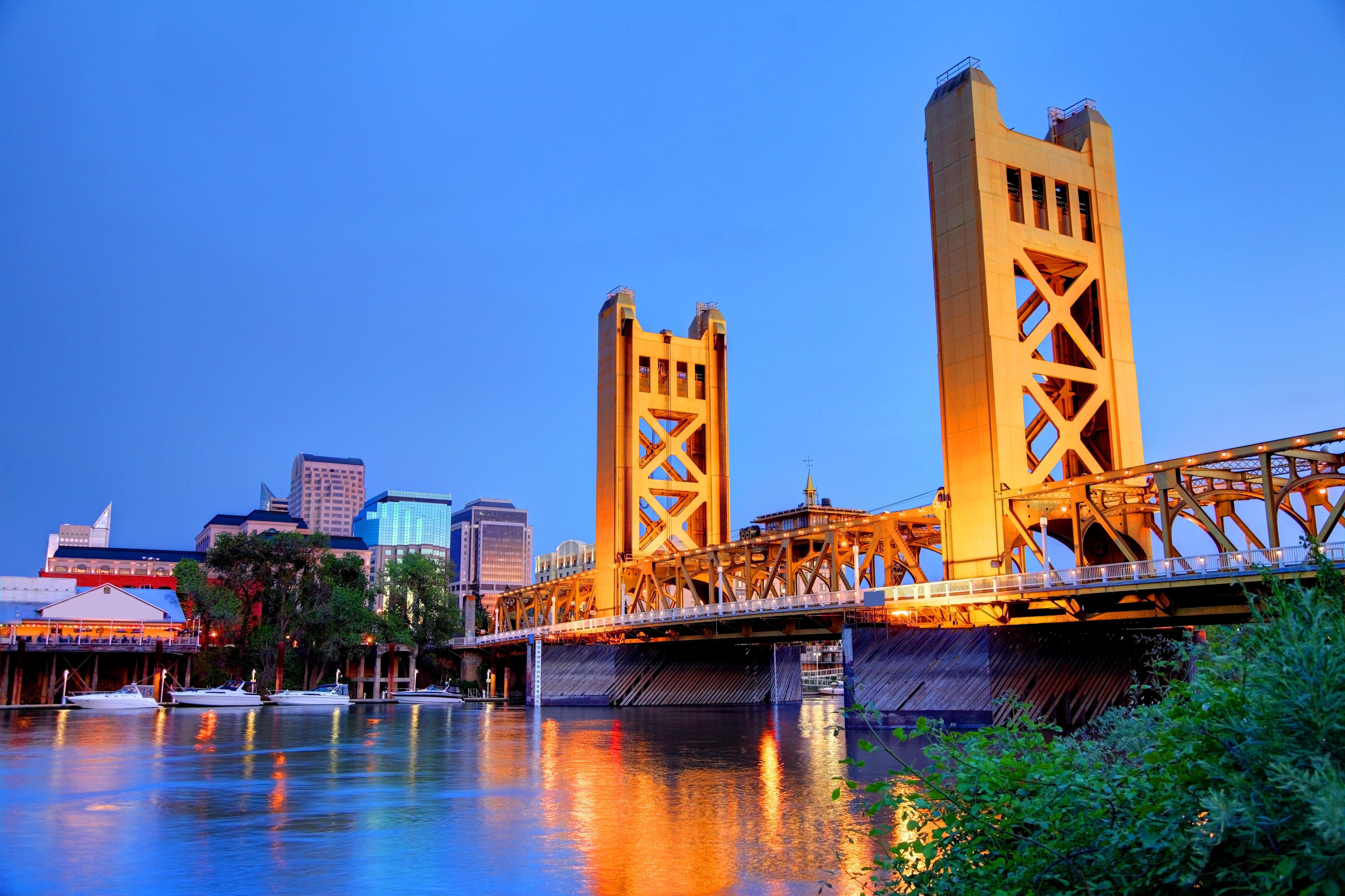 Colorful Sacramento skyline along the banks of the Sacramento River at night, with yellow tower bridge reflecting in the water, California