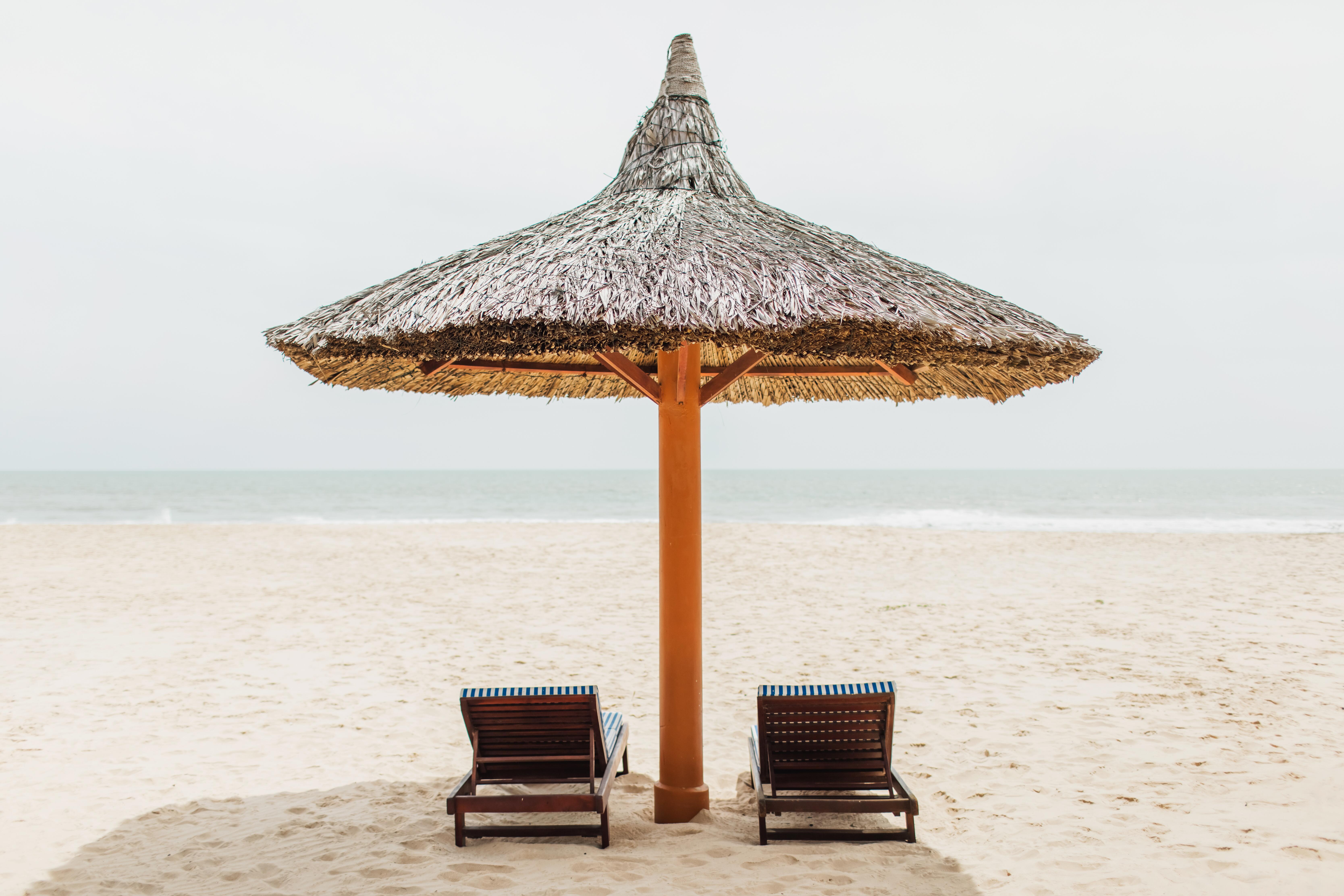 Image of a thatched/straw umbrella covering two chairs on a beach.