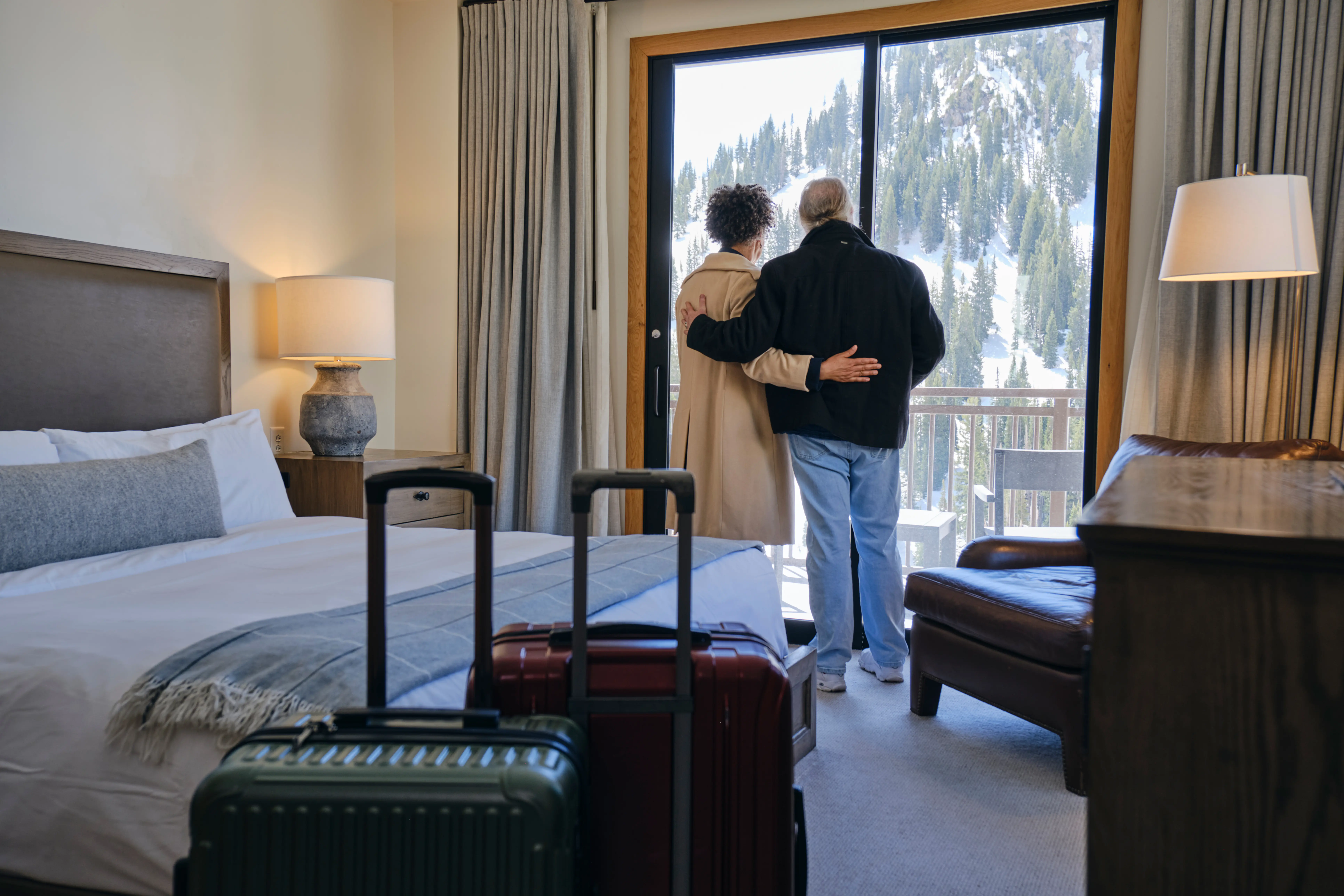 A senior aged couple entering their hotel room and enjoying the view of the snowy mountainside from their balcony doorway window at a ski resort