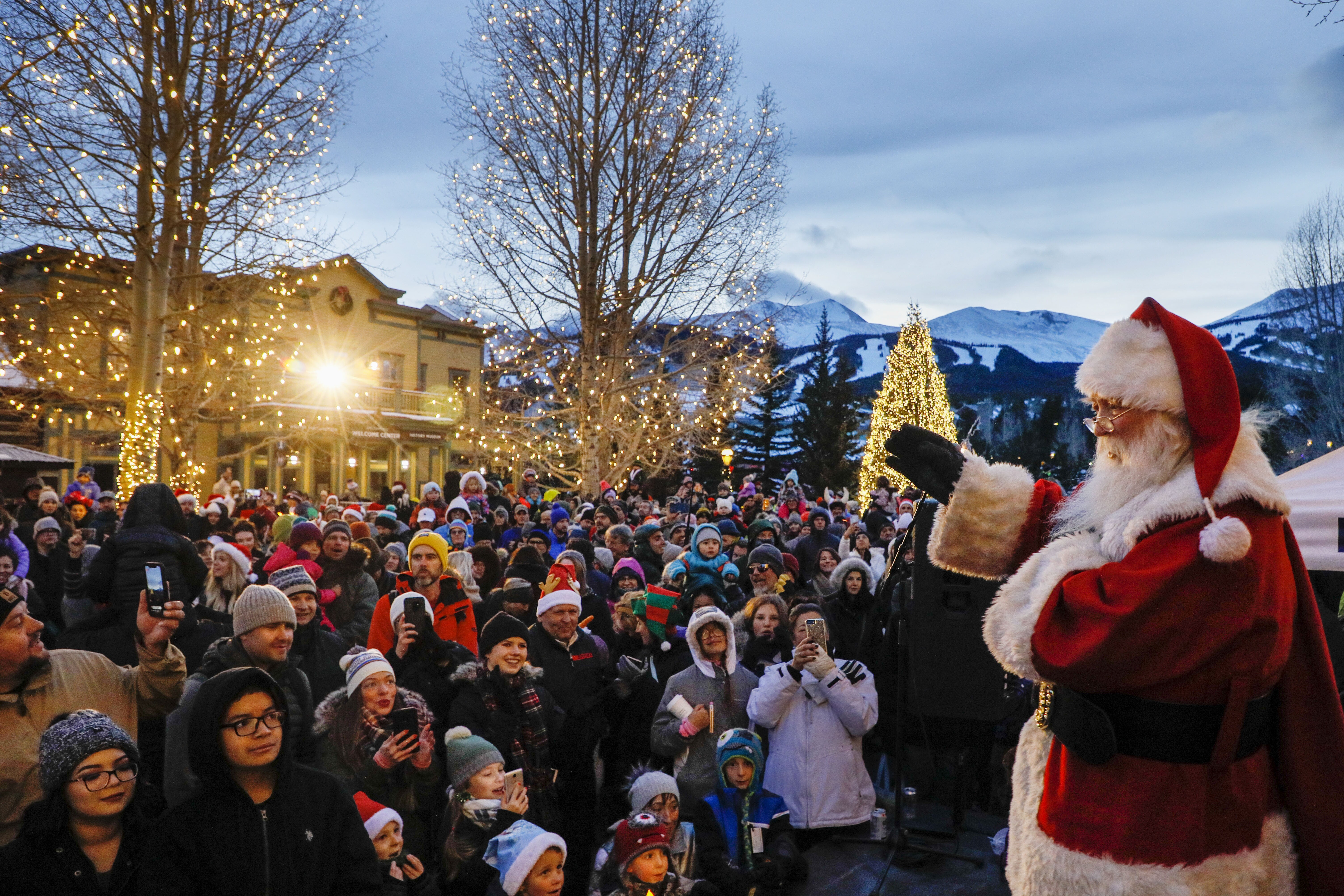Santa Speaks to crowd with snowy mountains and festively decorated store fronts in the background