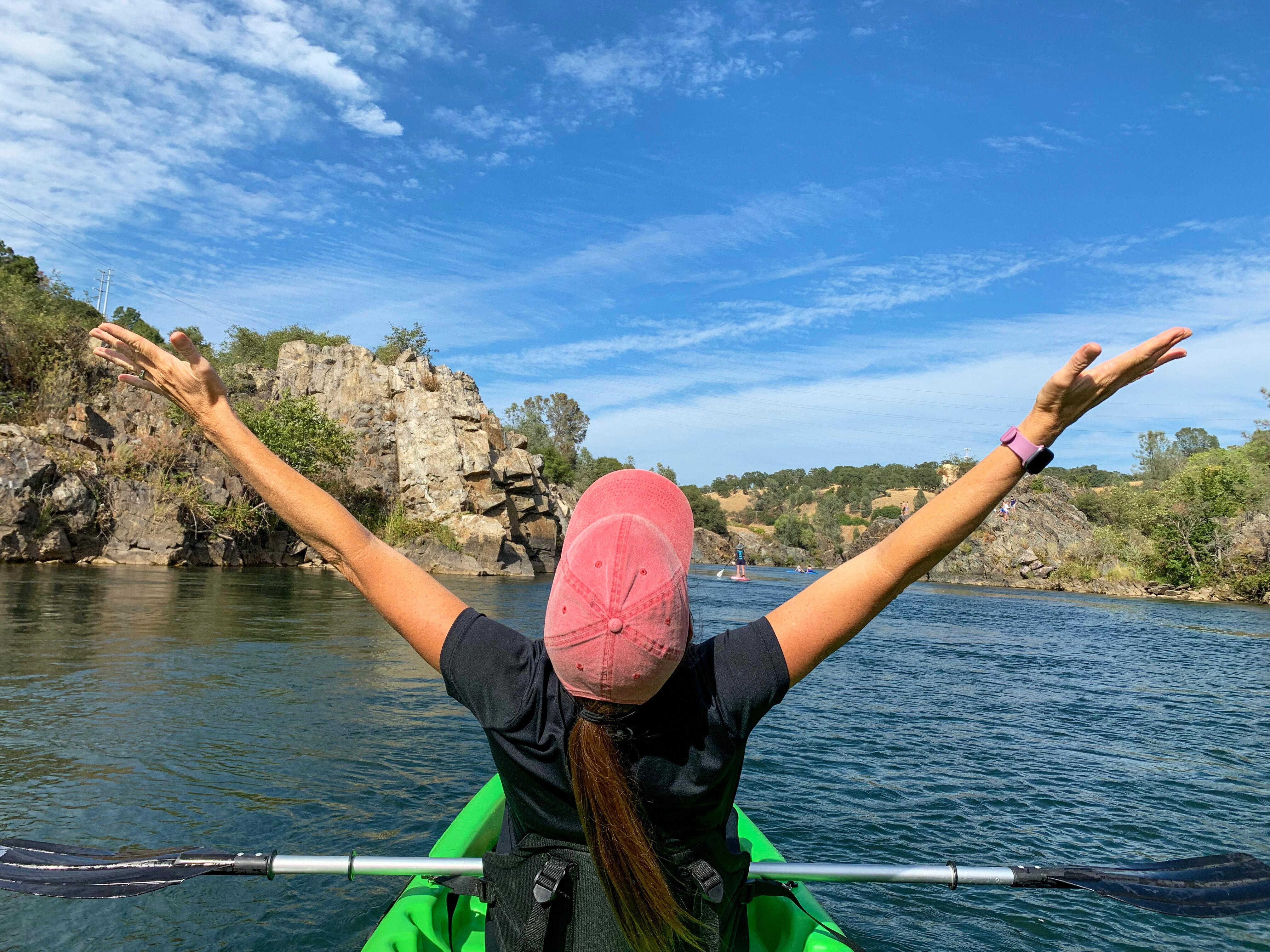 Point of view from behind a woman in green kayak with arms up celebrating a beautiful sunny day on Lake Notamas, with rock cliff shoreline, Falsom, California