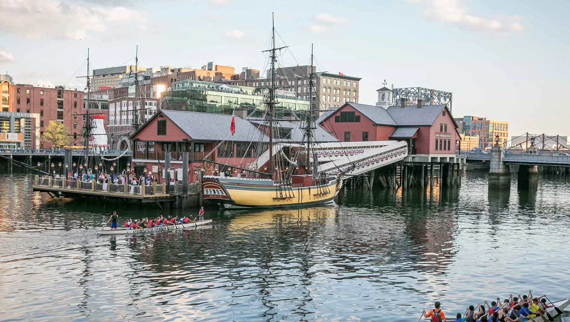 Old ships sitting docked in the Boston Harbor and people canoeing in the foreground.