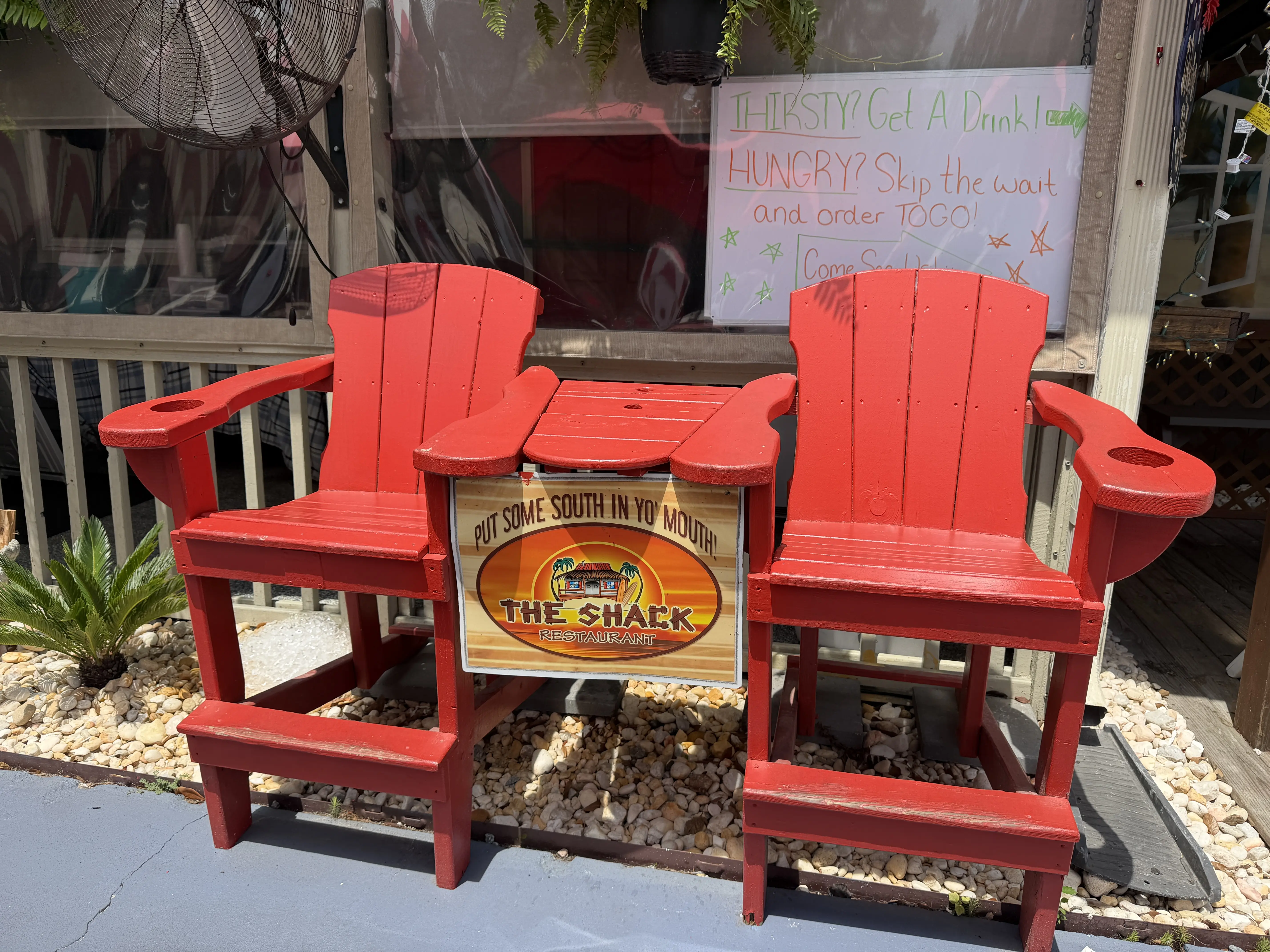 a set of red Adirondack chairs with a sign between them the reads " Put some South in yo' mouth! The Shack. Bove the chairs is a white board with specials on it.