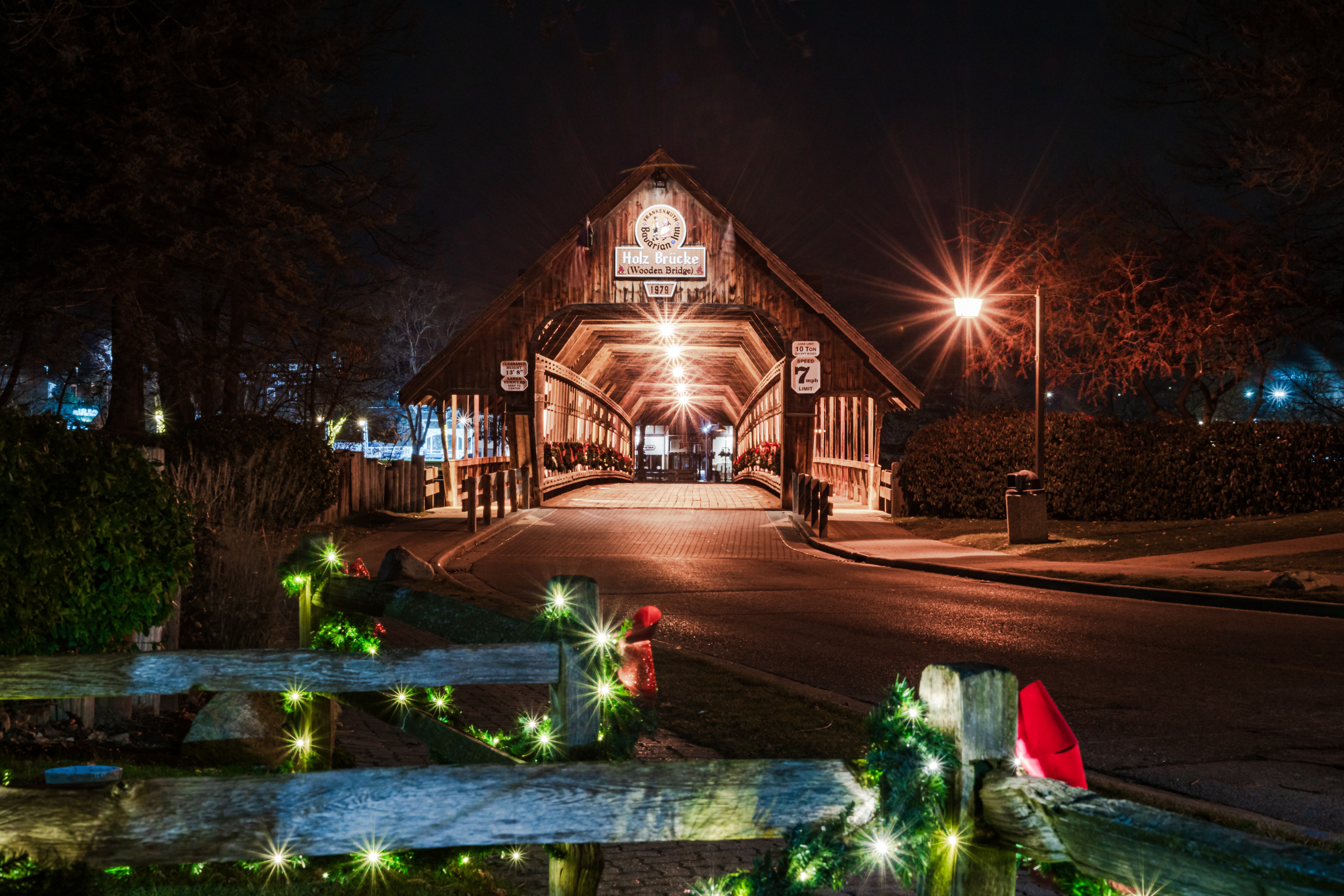 Wooden covered bridge lit at night with holiday garland draped over fencing in foreground