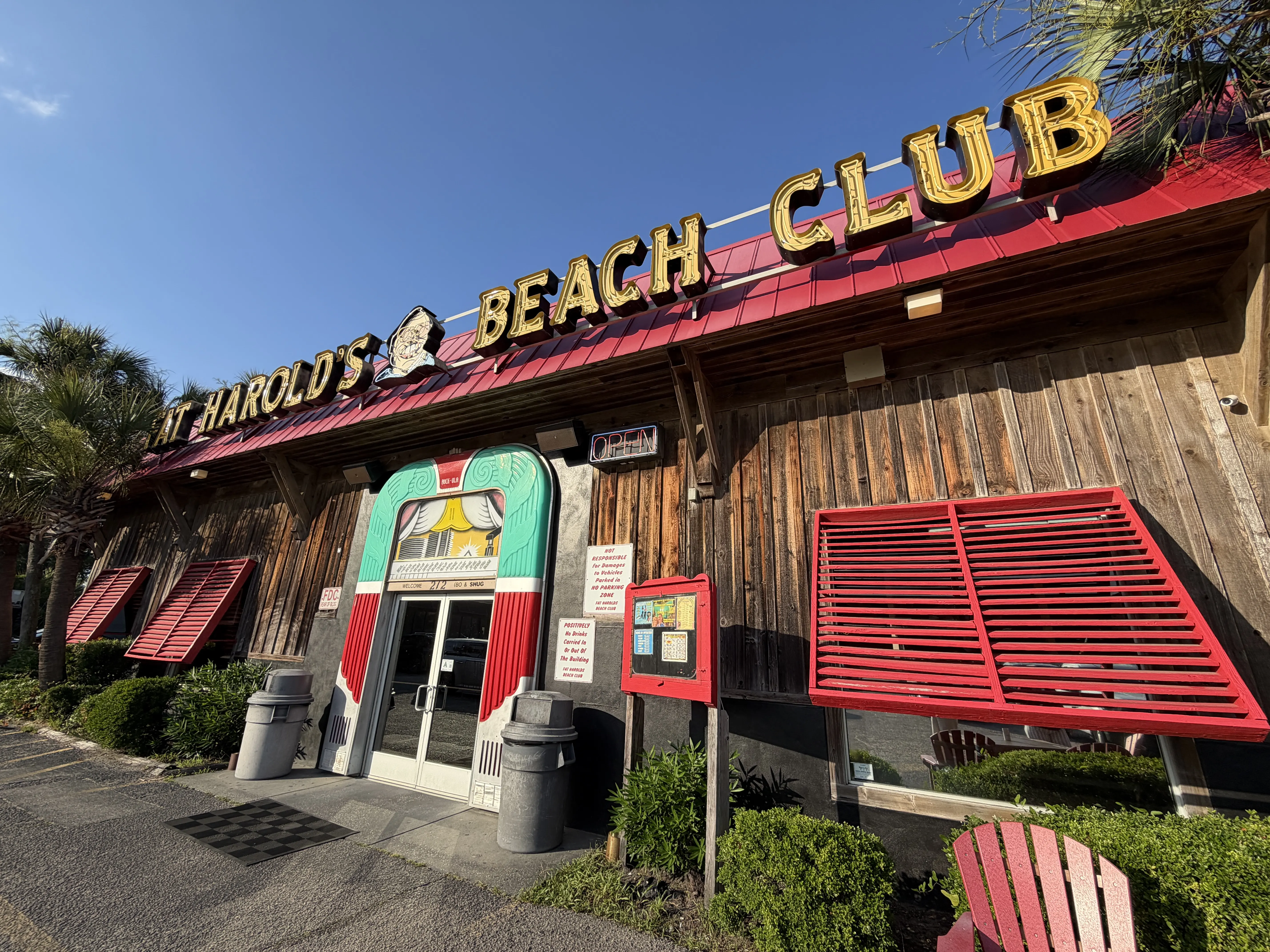 Exterior of dark wood clad building with yellow neon letters on a red roof that reads "Fat Harold's Beach Club. Windows have red shutters.