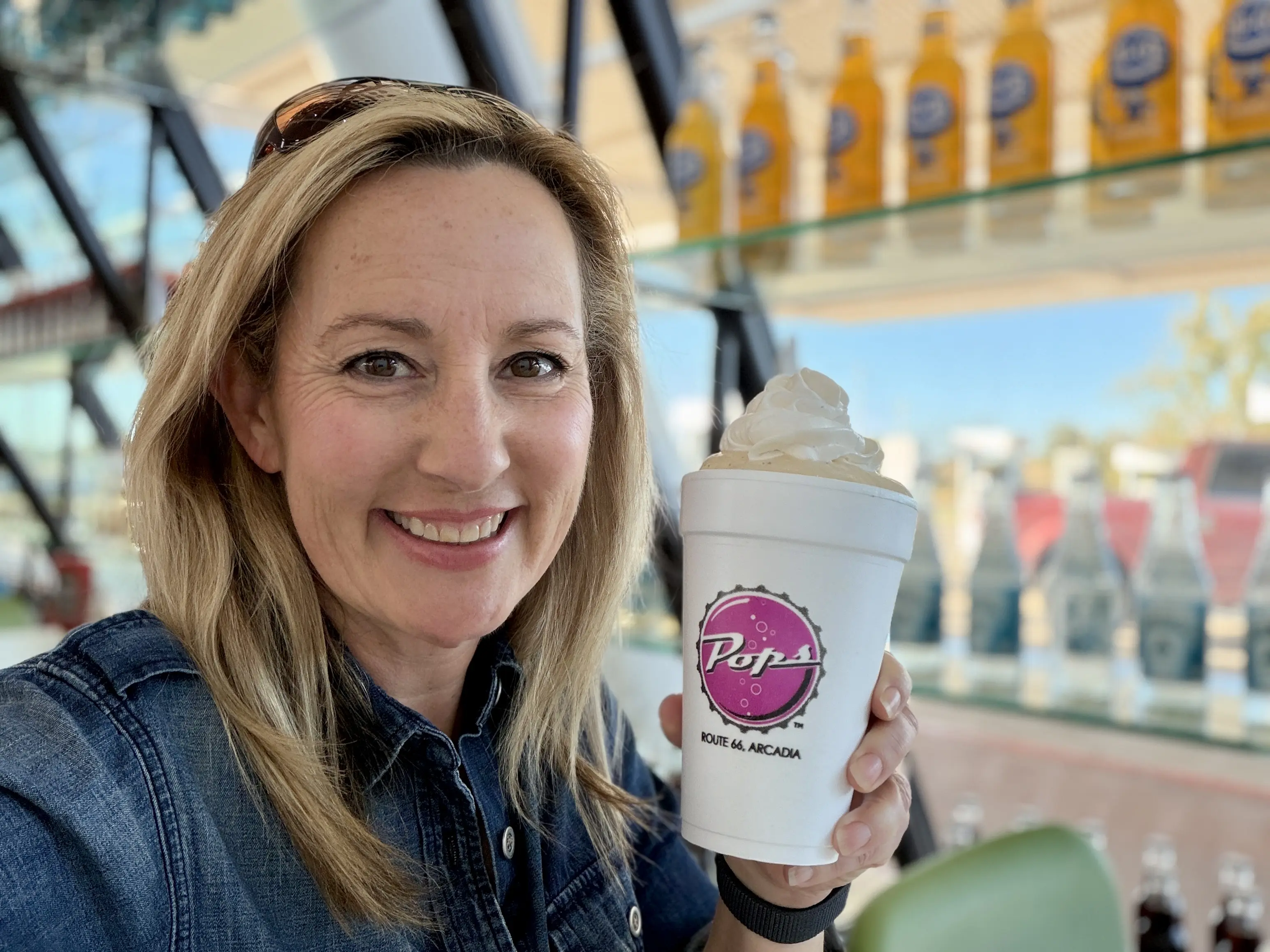 Woman with blonde hair holds up white cup with Pops branding that is filled with a milkshake topped with whipped cream