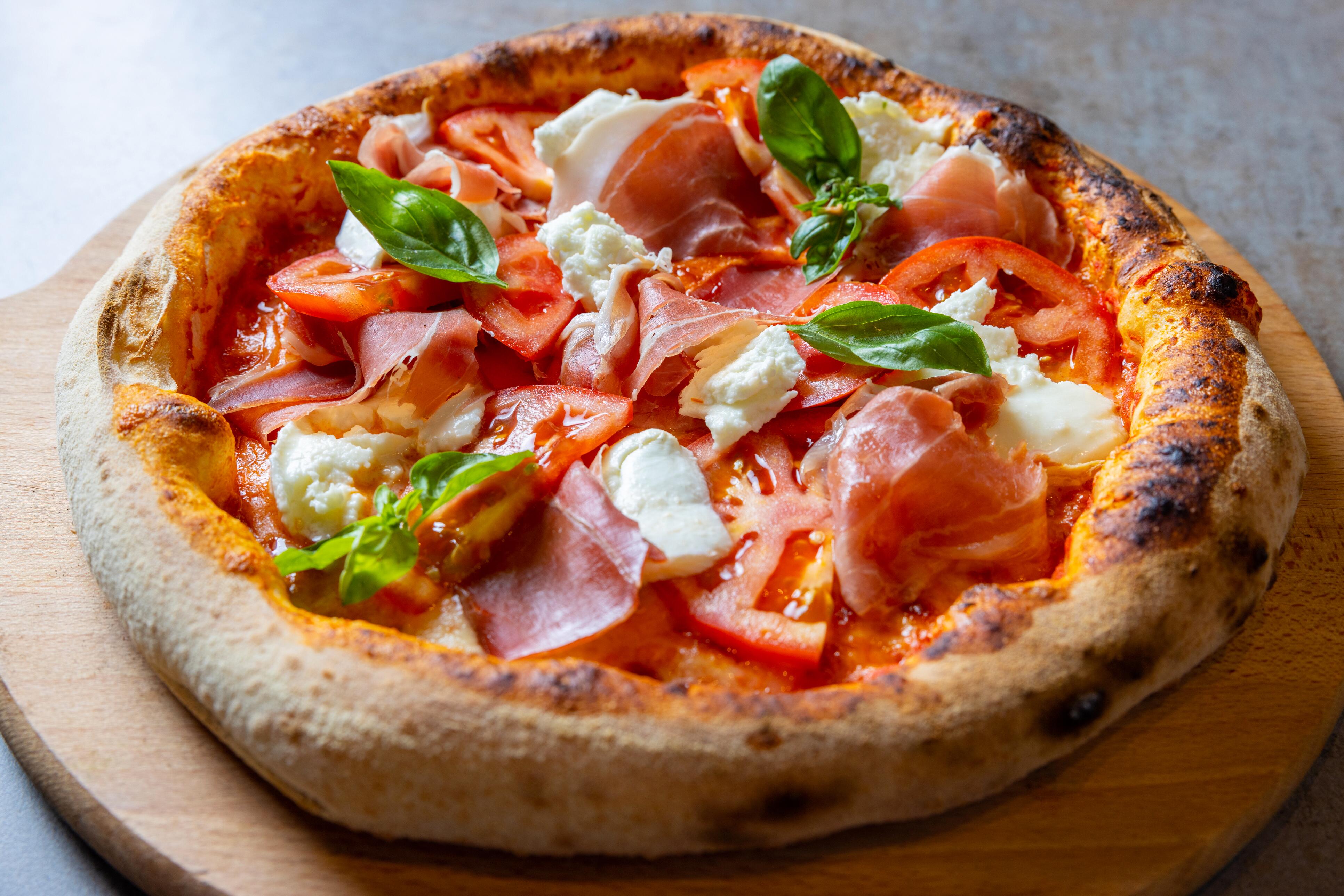 Woodfired pizza with prosciutto, mozzarella, tomato slices, and basil leaves