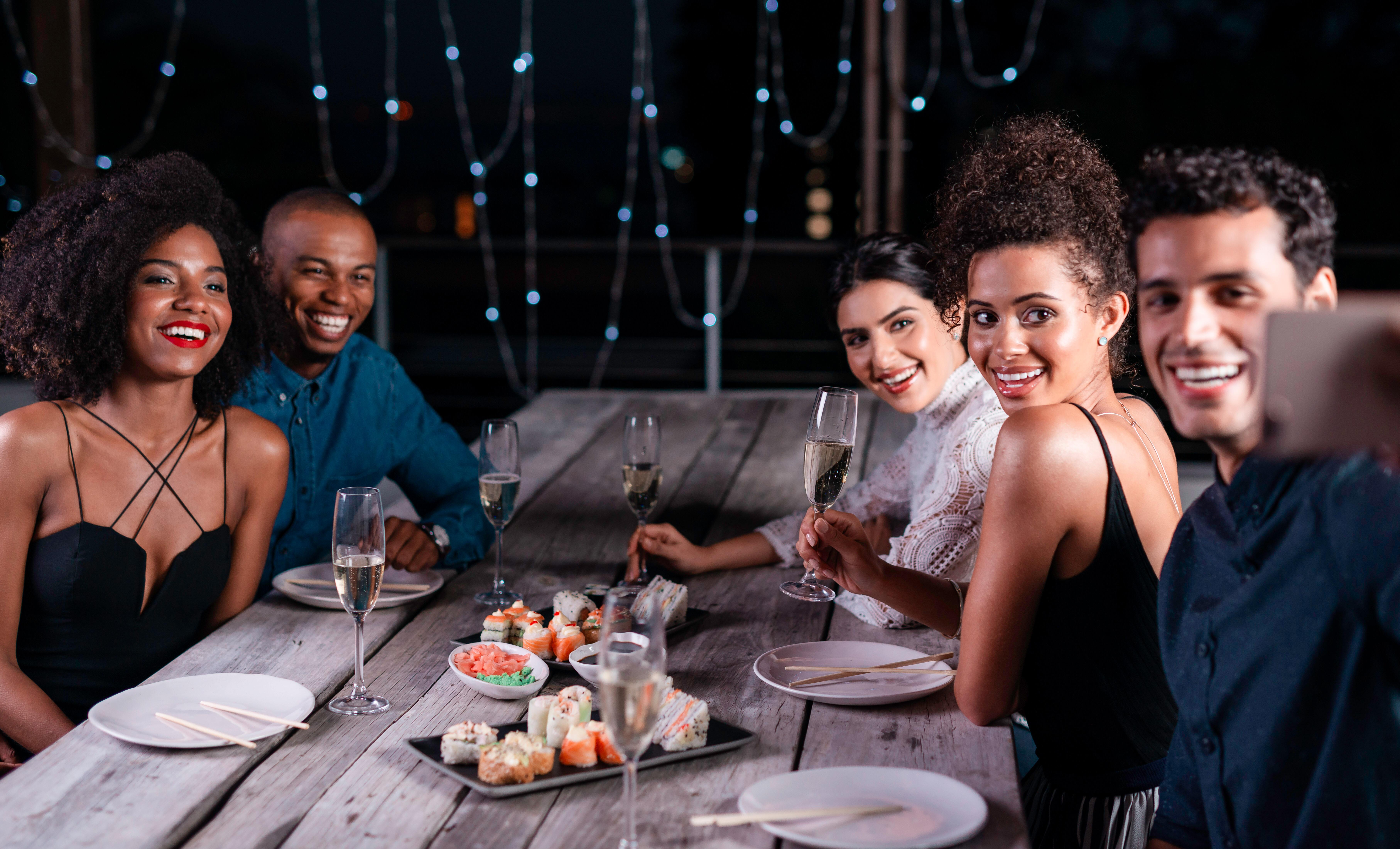 A group of friends seated at a wooden table for an elegant outdoor dinner party, sharing sushi and raising glasses of sparkling wine. The atmosphere is lively and joyful, enhanced by string lights and an intimate setting.