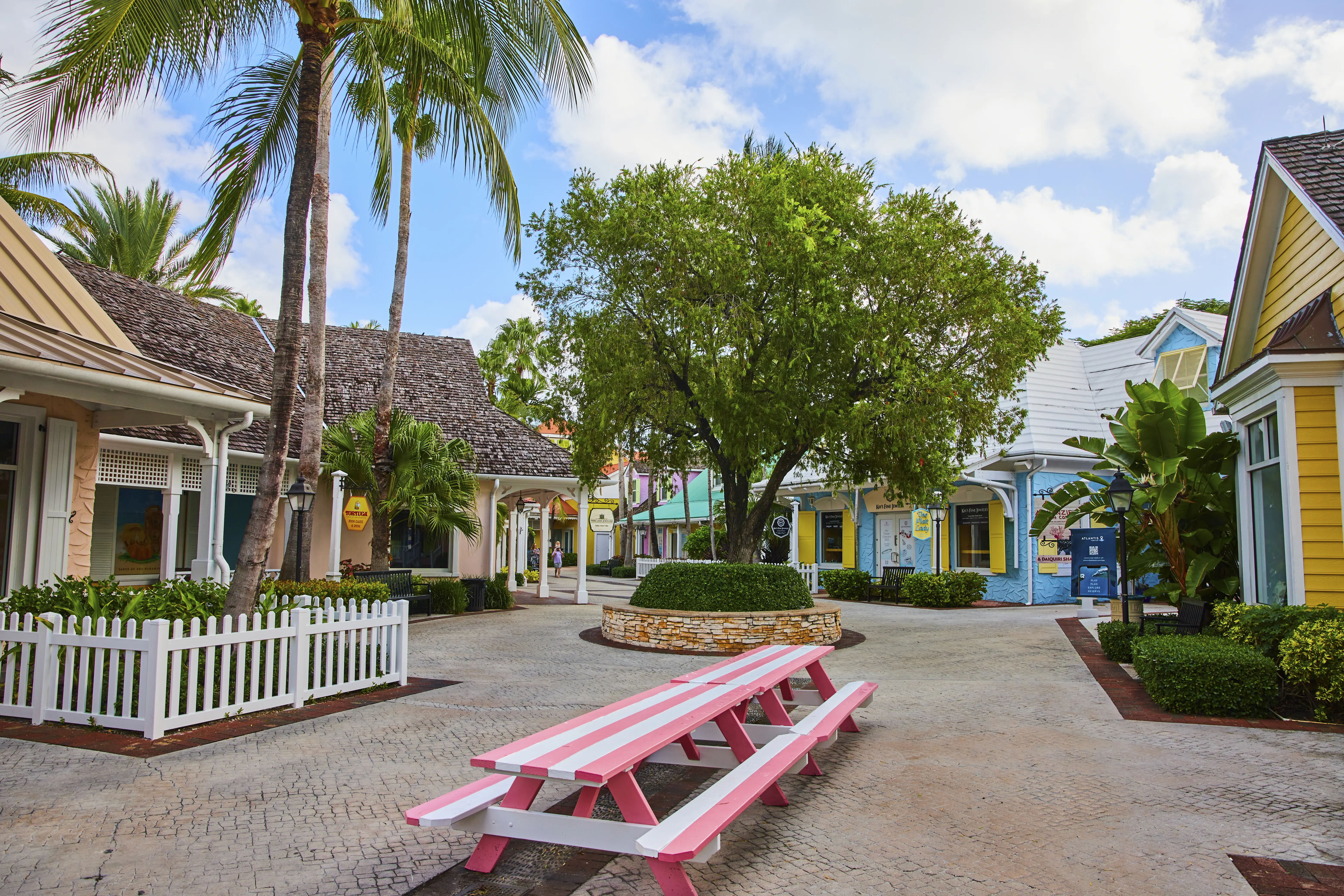 Vibrant tropical shopping village in Nassau, Bahamas, featuring pastel colored buildings, a pink picnic table, and lush palm trees.