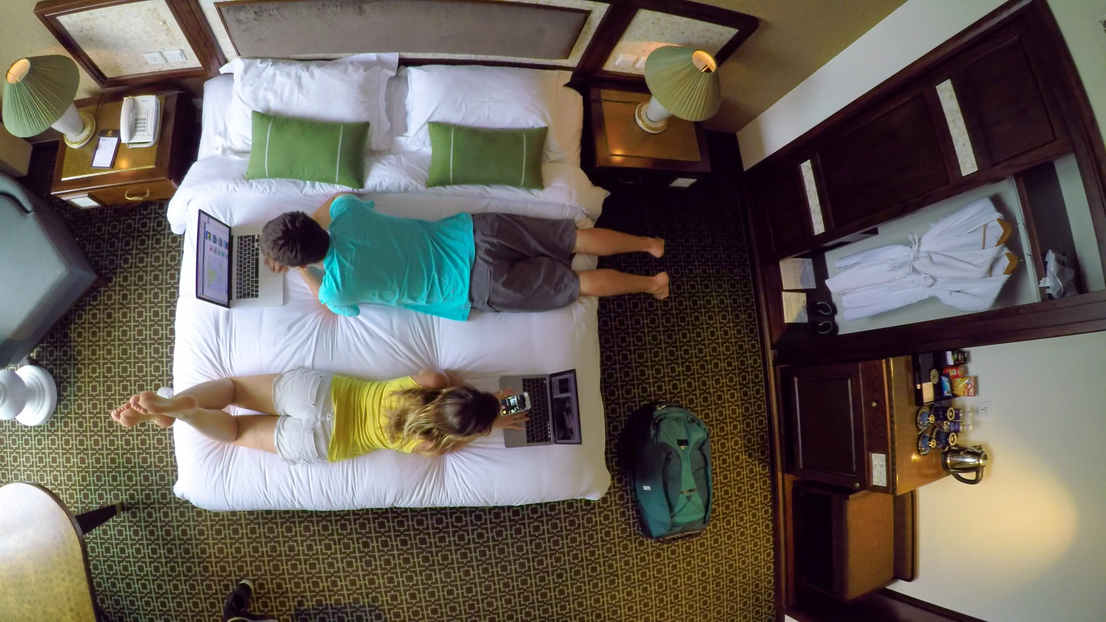 Male and female digital nomads working on computers from their hotel room while lying on the comfortable king size bed. Young tourist couple surfing the internet in their holiday suite.
